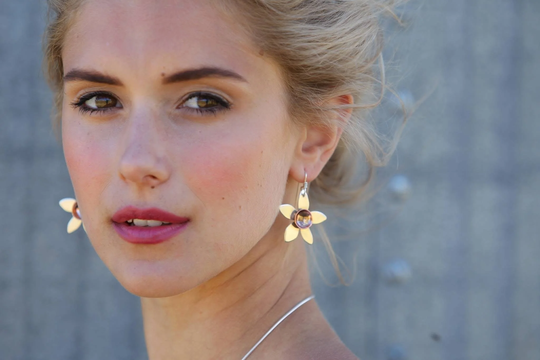 Close-up of a woman's face with gold floral earrings, light makeup, and blonde hair styled in waves, outdoors with a blurred background.
