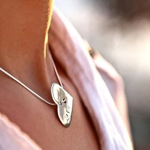Close-up of a woman wearing a silver heart-shaped necklace with engraved initials and a small infinity symbol.