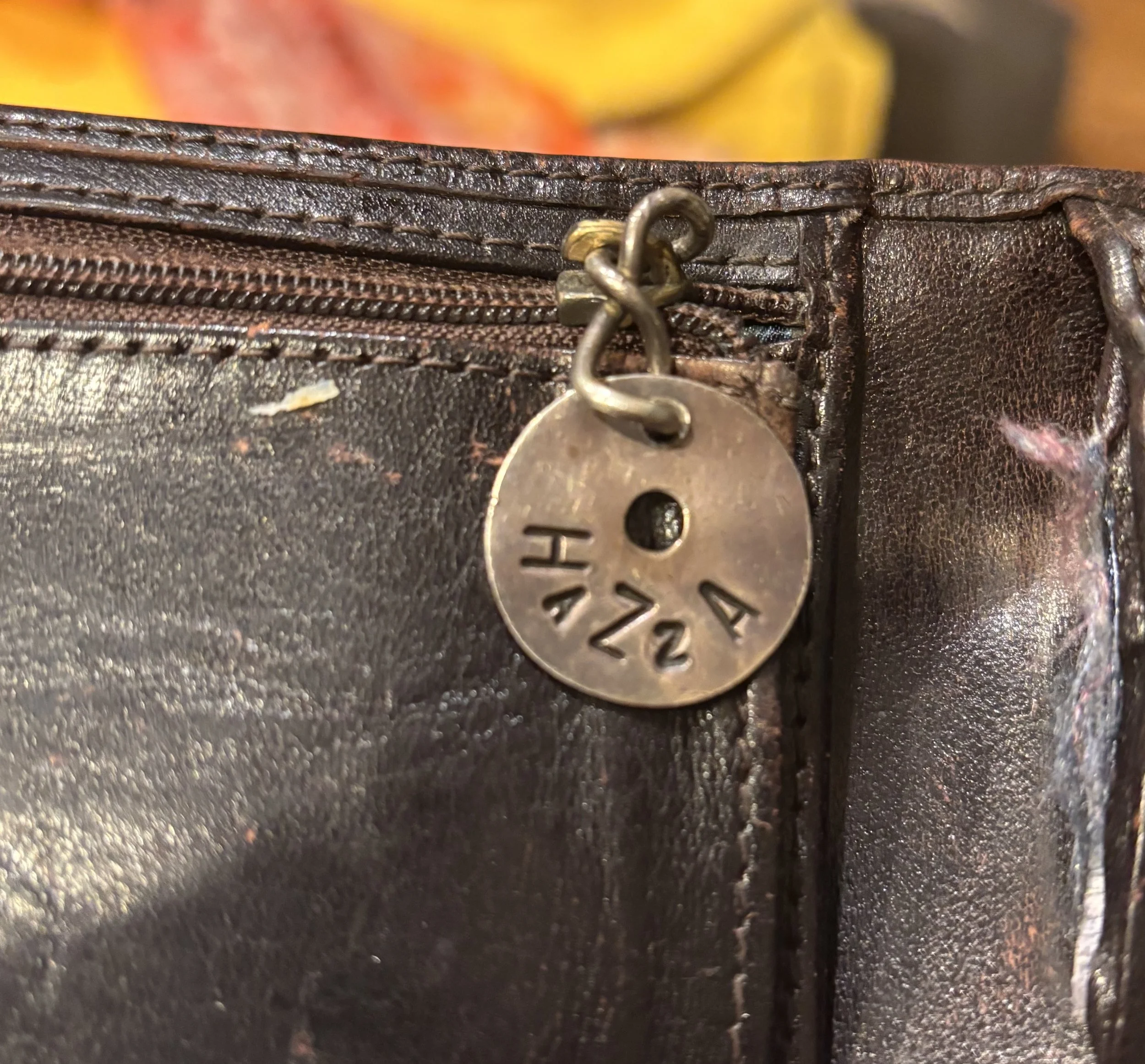 Close-up photo of a brown leather wallet with a metal zipper and a metal keychain with a circular tag attached, showing visible stitches and a textured surface.