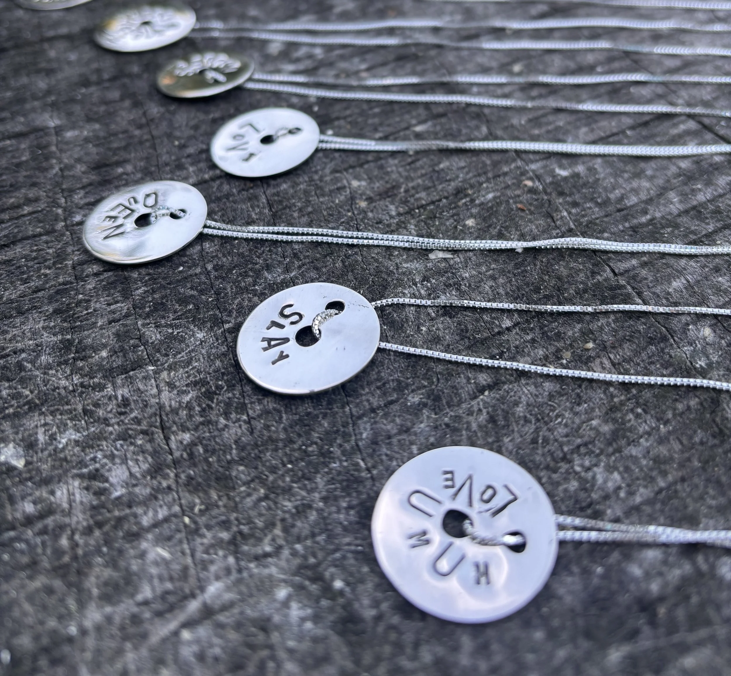 Close-up of several silver necklaces with circular, engraved pendants laid out on a rough, wooden surface.