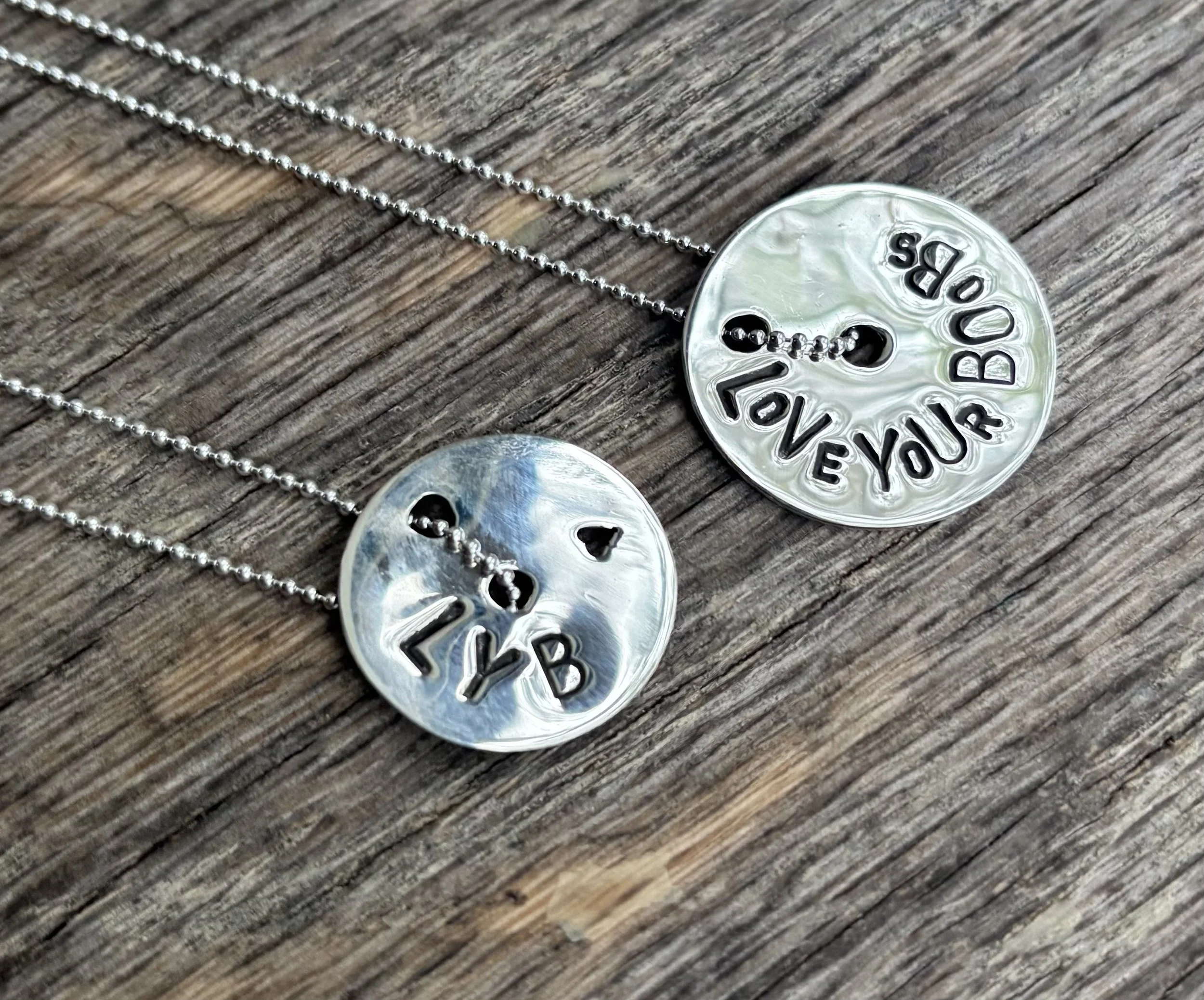 Close-up of a silver necklace with a pendant featuring a cut-out smiley face and the words 'You Are So Loved' on a wooden surface.