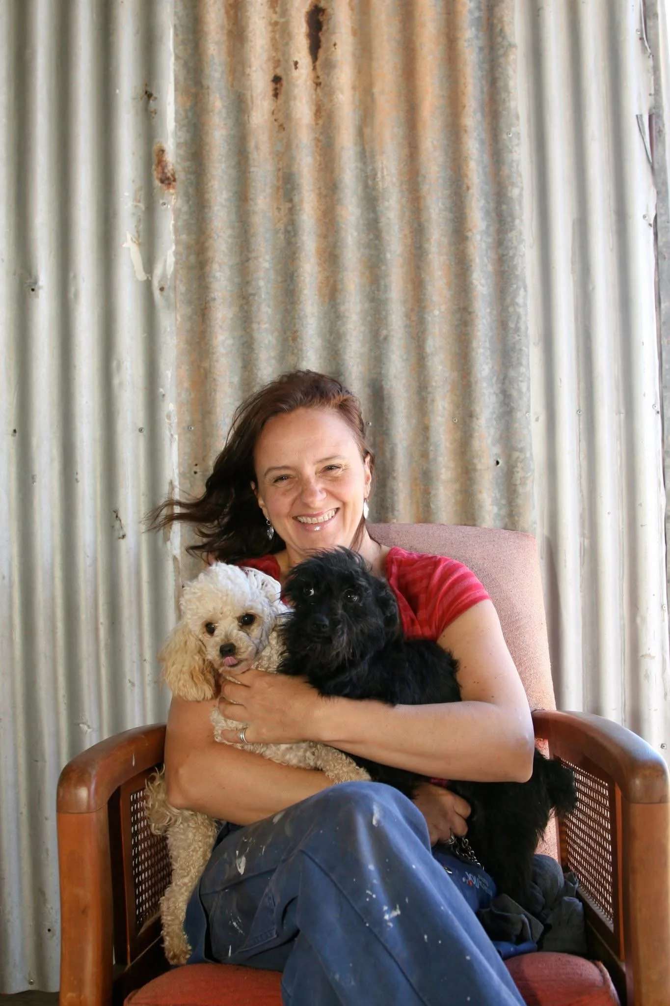 A woman sitting on a wooden chair, holding three dogs, while smiling at the camera. The background features corrugated metal sheets.