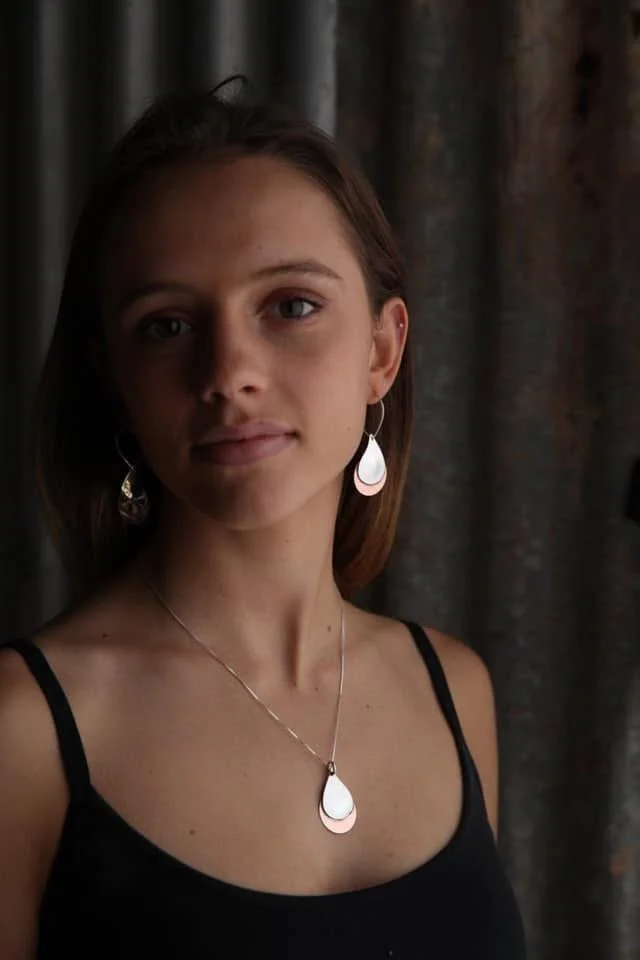 A young woman with brown hair and blue eyes wearing teardrop-shaped earrings and a matching necklace, dressed in a black tank top, standing in front of a dark curtain.