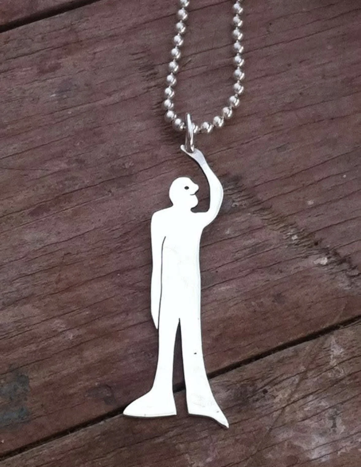 A silver necklace with a pendant shaped like a person with long hair. The pendant shows only the outline of the figure with a raised arm, which the necklace chain is attached to. The background is a wooden surface.