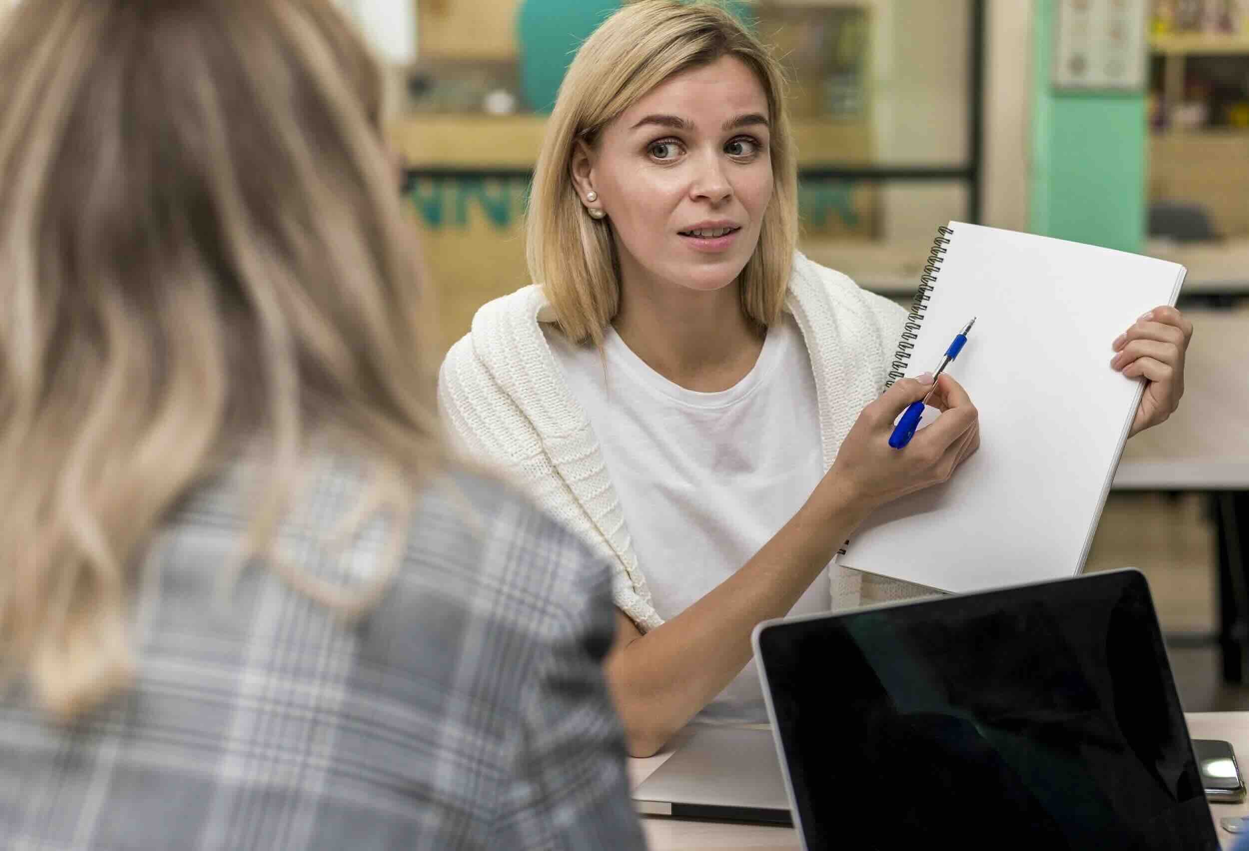 Woman providing executive function coaching to a client, pointing to notes on a pad in a bright classroom setting.