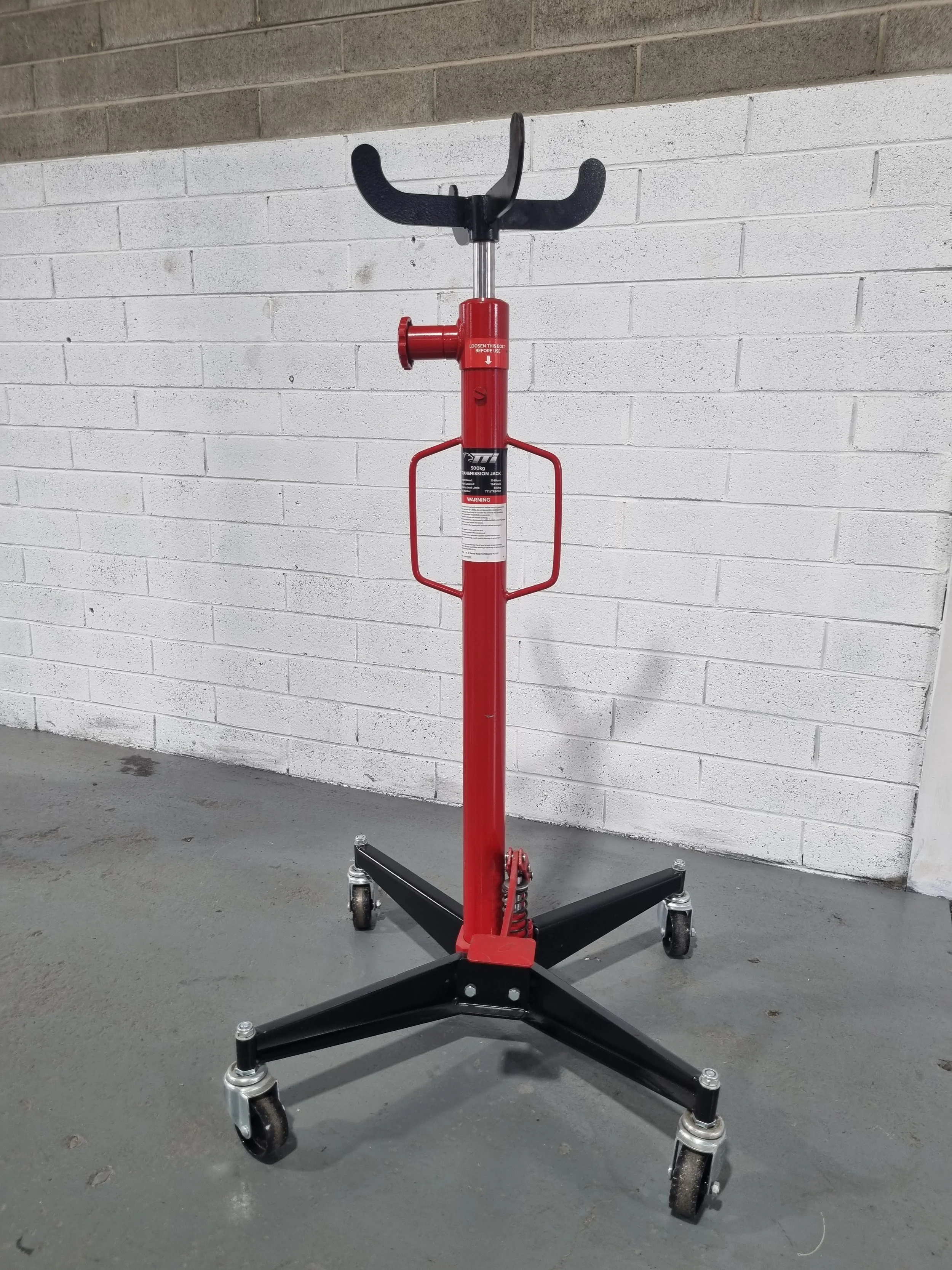 A red hydraulic moving dolly with black handles and wheels, positioned on a gray concrete floor against a white brick wall.