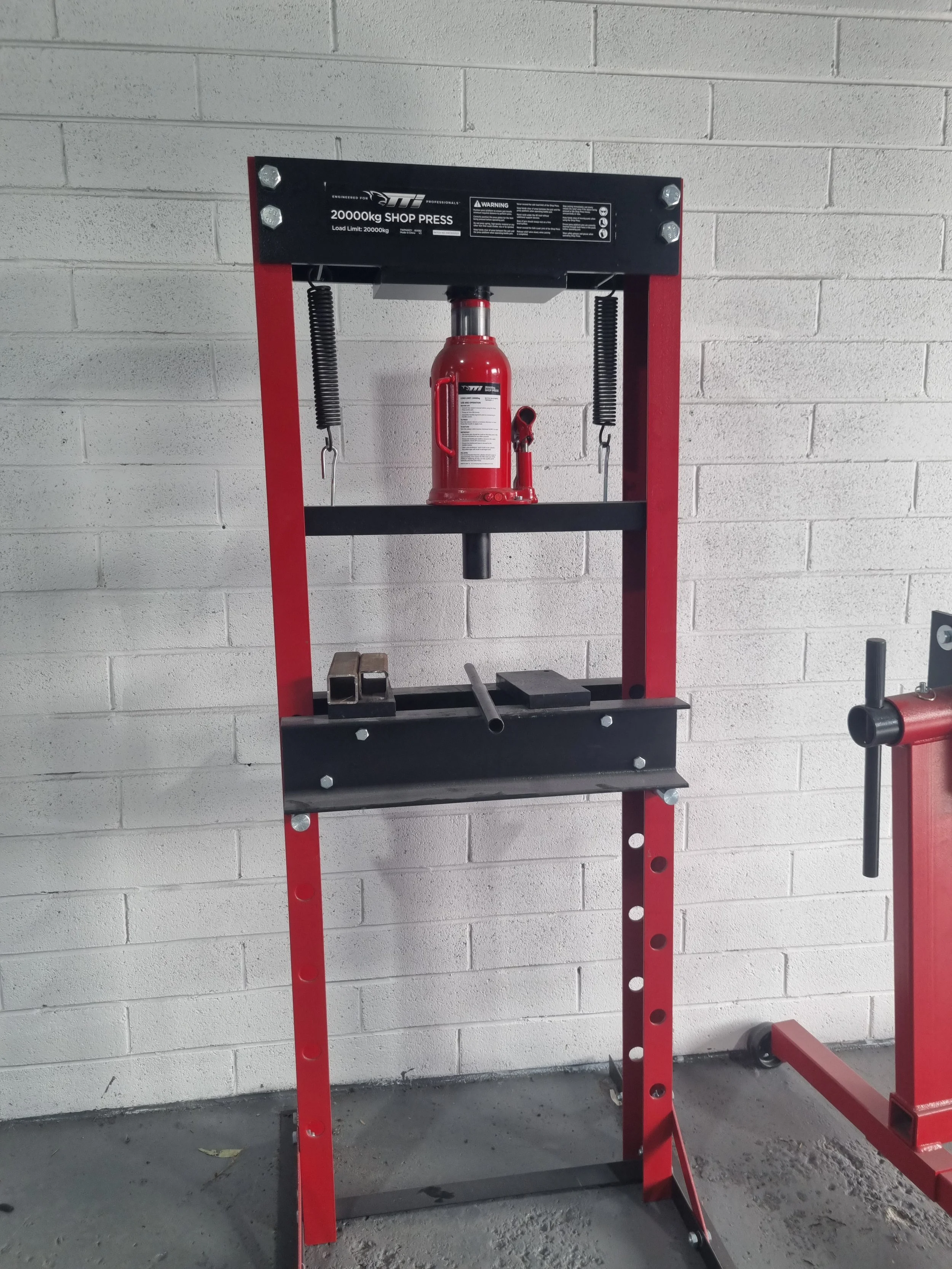 A red hydraulic shop press with a fire extinguisher attached to the center, set against a white brick wall.
