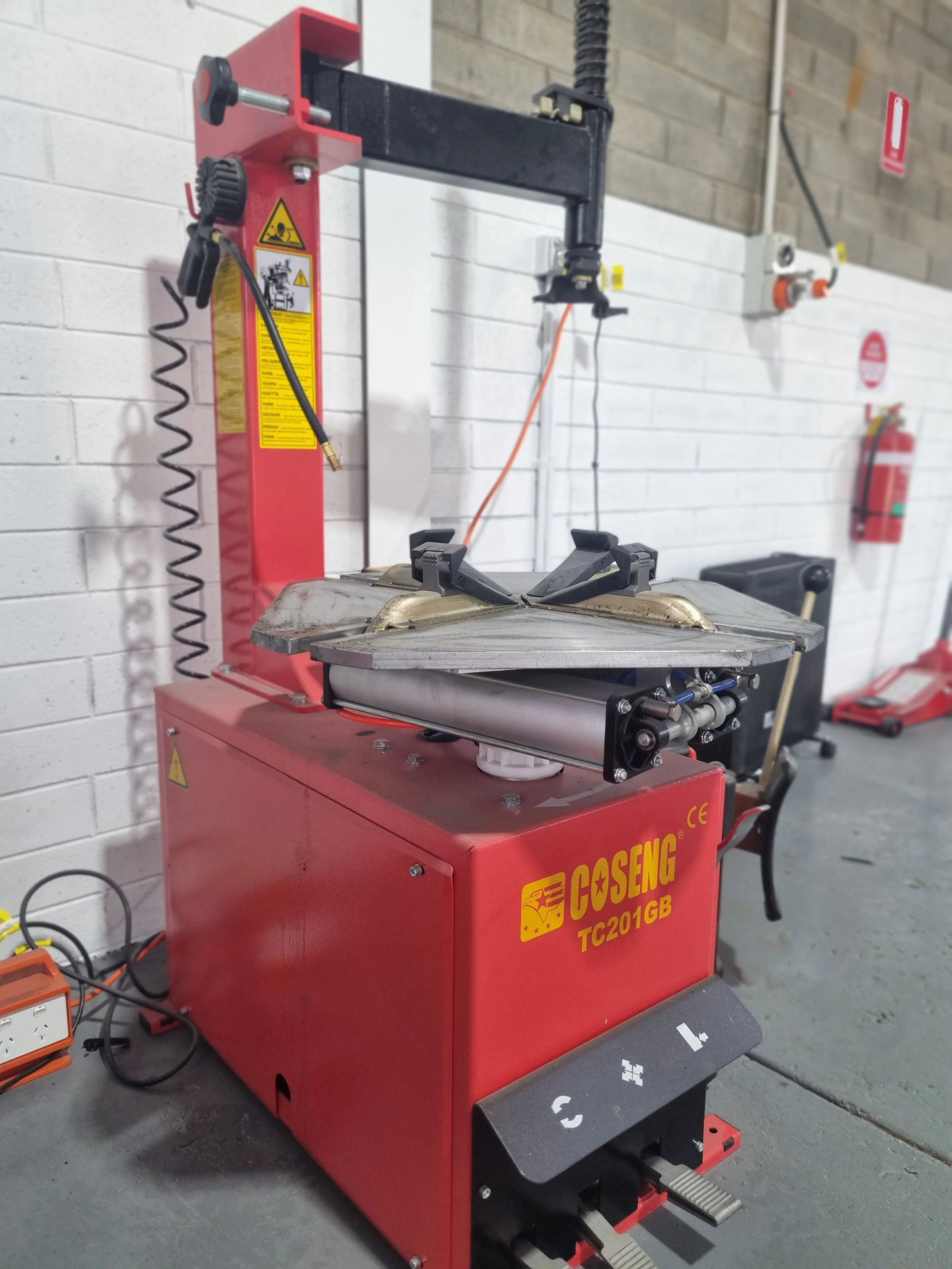 A tire curing machine in a workshop. It is red and labeled 'COSENG TC201GB'. The machine has clamps holding a tire mold, with various controls and a foot pedal for operation.