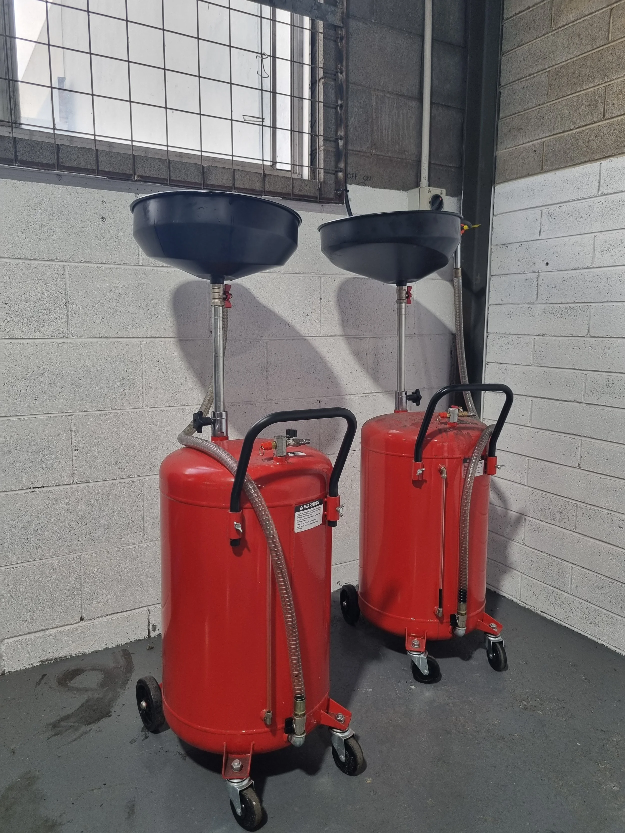 Two red oil drain tanks on wheels in a garage, with black funnel shaped tops and tubing attached.