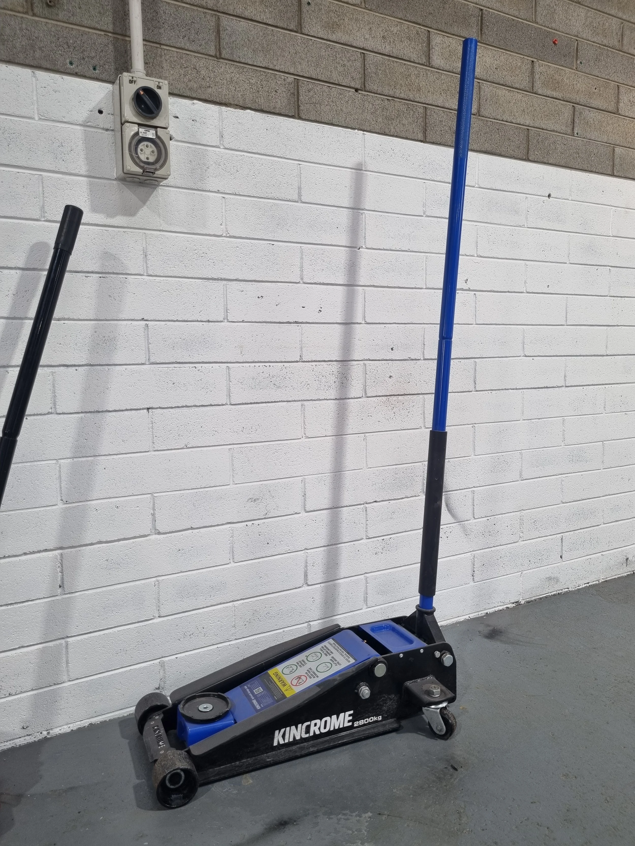 Blue and black hydraulic floor jack resting on the gray concrete floor, with a white brick wall in the background and a blue broom leaning against the wall.