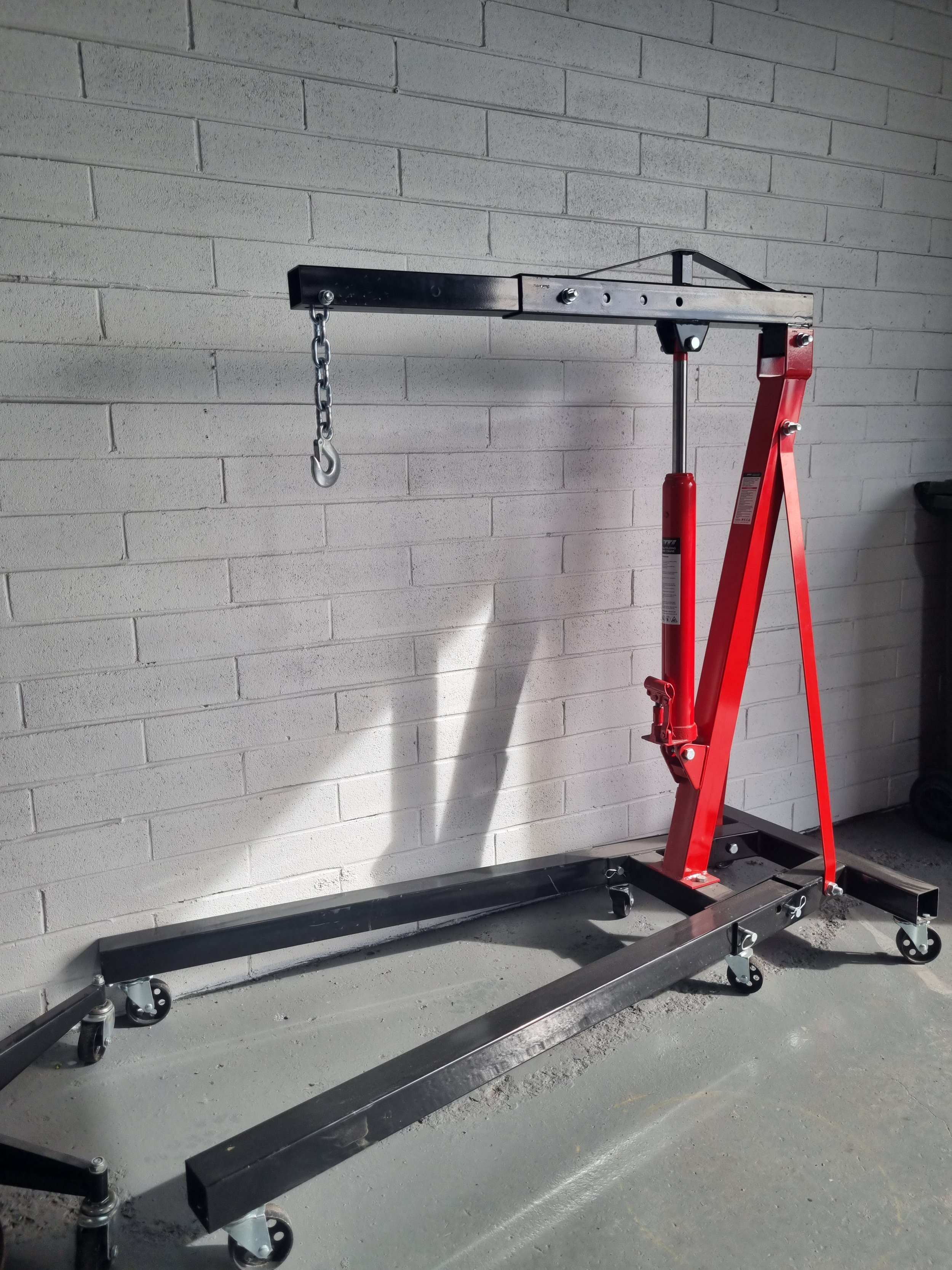 A red and black engine hoist with a long metal arm and a chain hanging from it, on a concrete floor next to a brick wall.