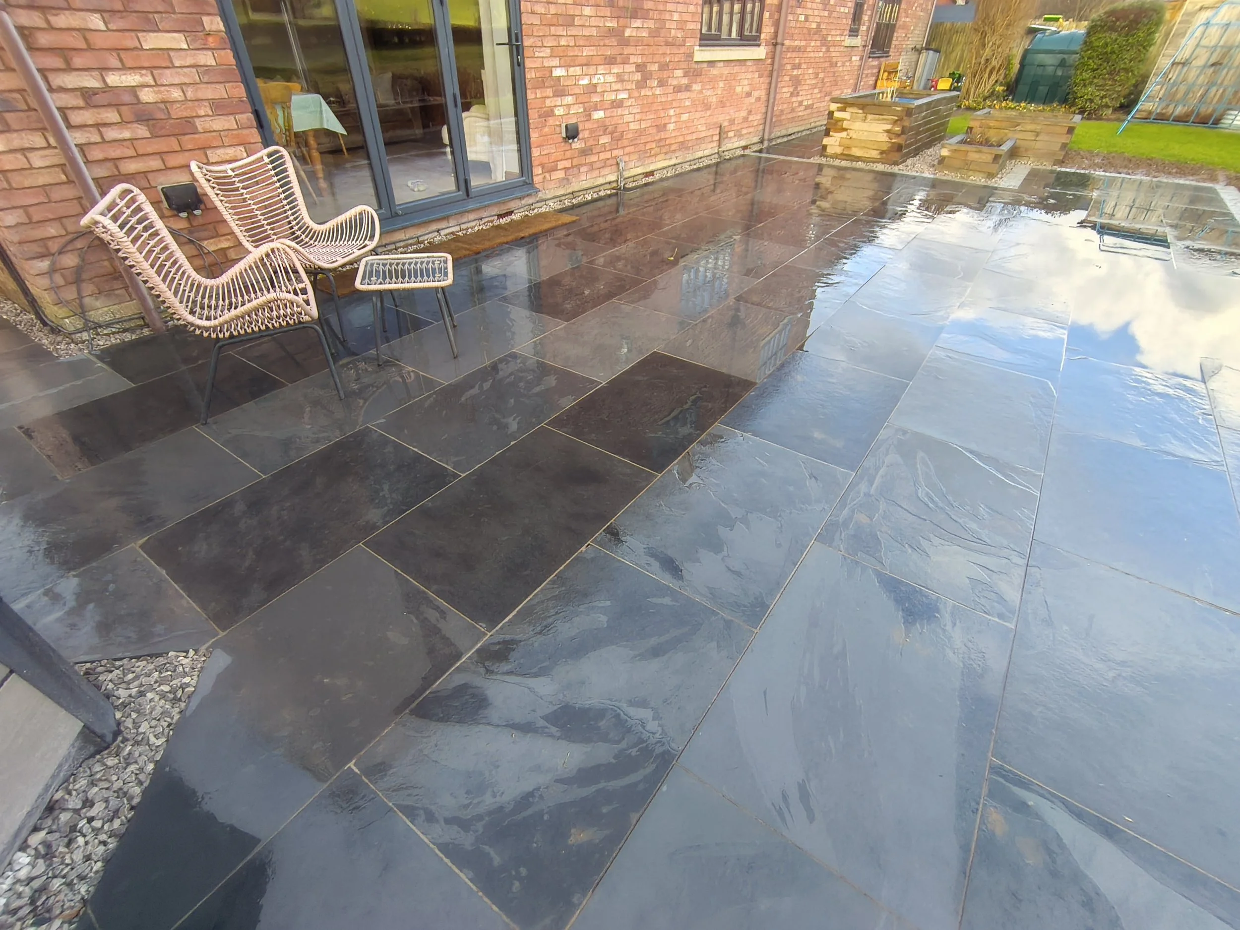 Wet outdoor patio area with black and dark brown stone tiles, some reflecting the sky and clouds. Two wicker chairs with black metal frames and matching small table sit near a brick house with sliding glass doors. There are stacked wooden pallets and a small garden with green grass and plants in the background.