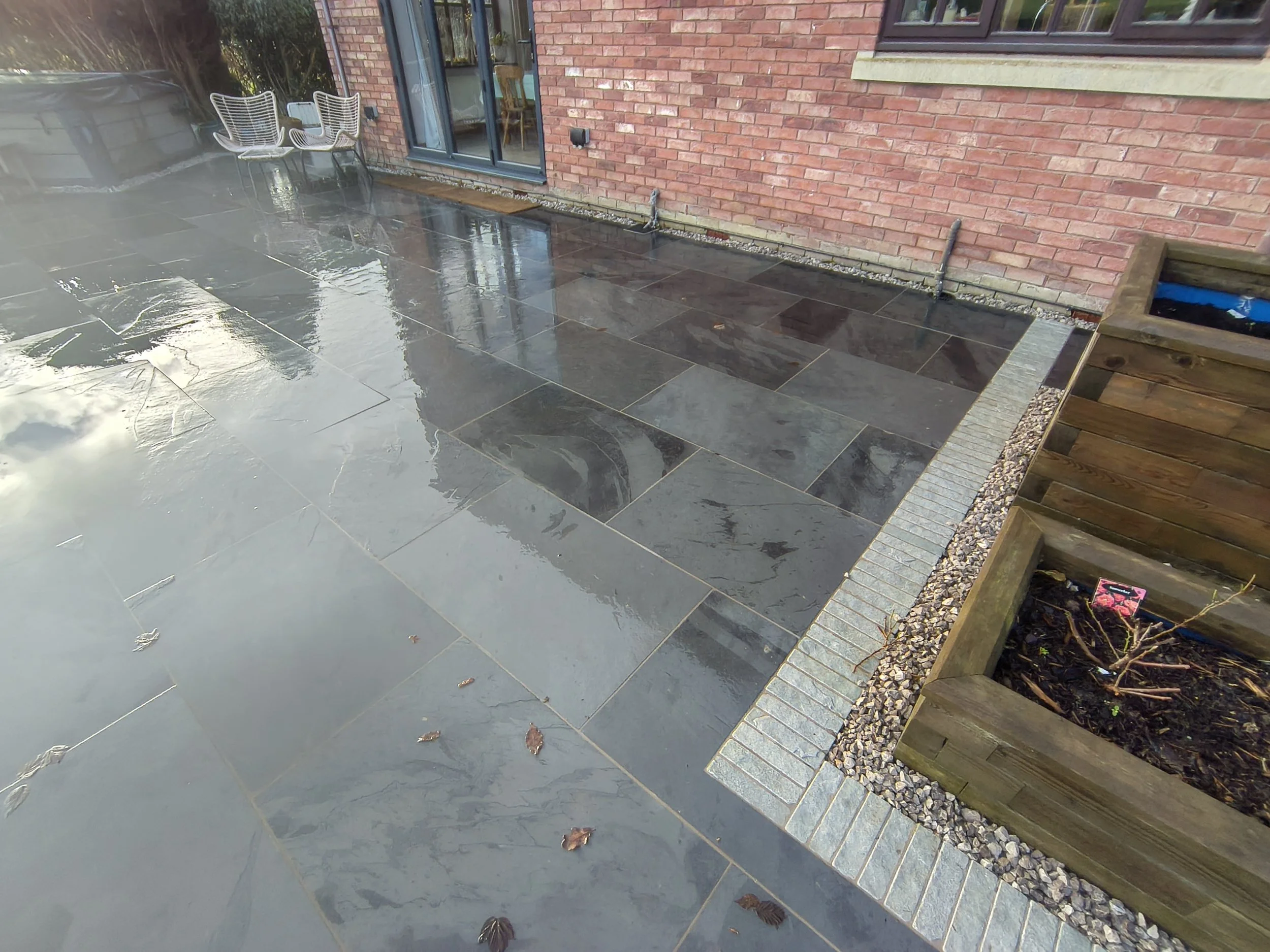 Wet black stone patio outside a brick house with sliding glass doors. There are two white outdoor chairs near the house. To the right, there is a wooden raised garden bed with soil and a pink plant marker.