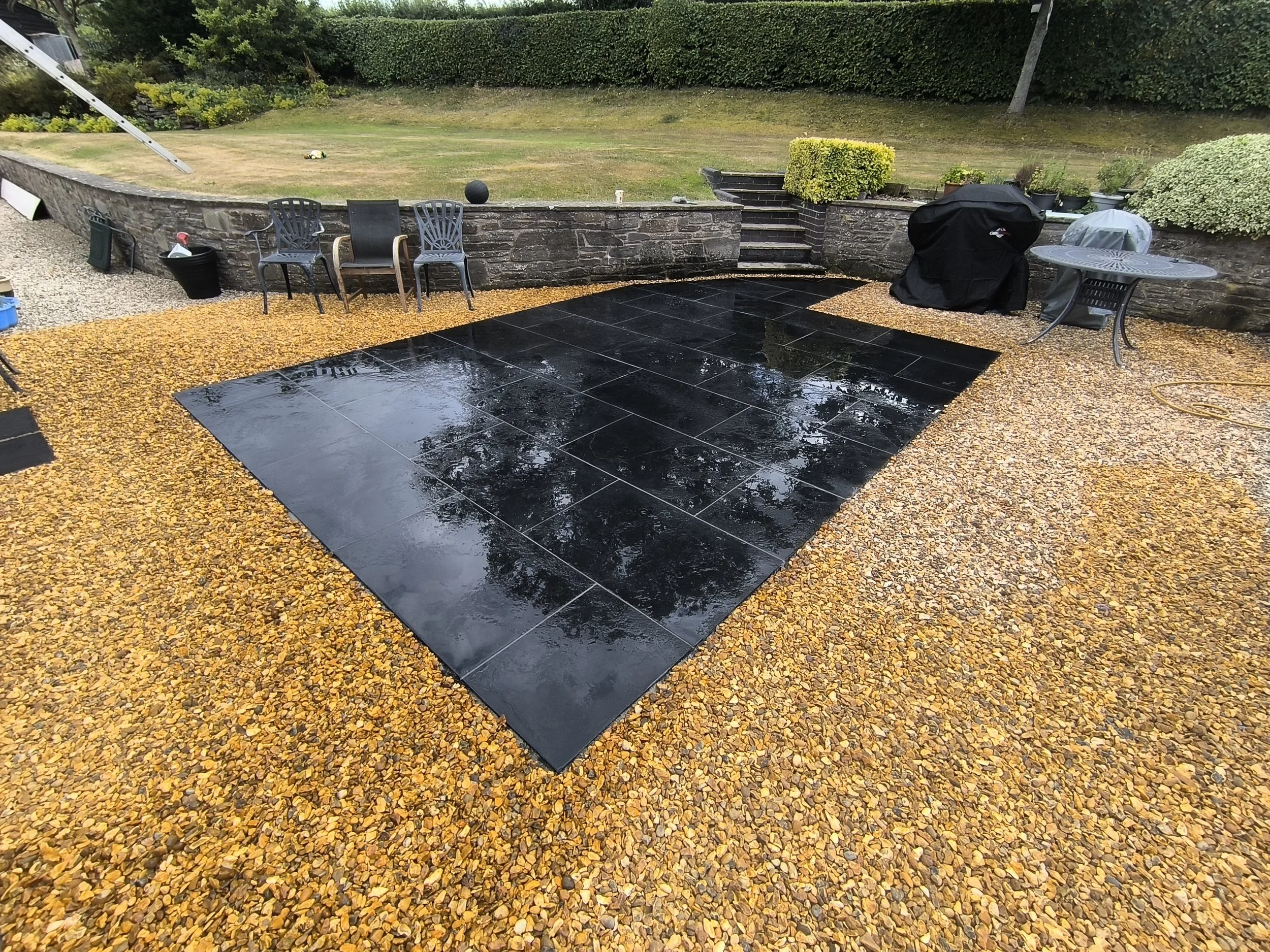 A backyard patio with black polished tiles, wet from rain, surrounded by a yellow gravel area, with chairs, a table, and a barbecue grill covered with a black cover, overcast weather.