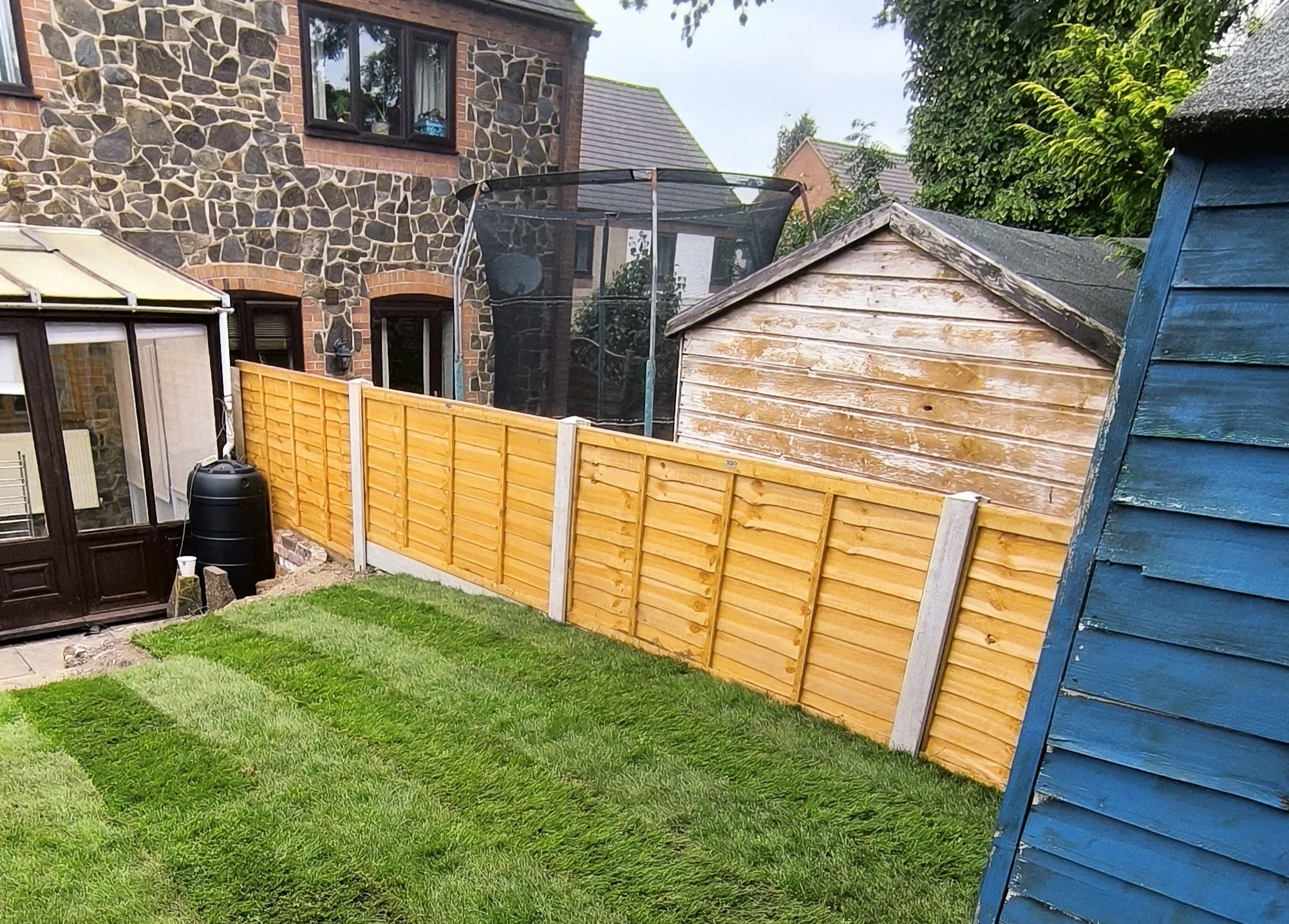 View of a backyard with a freshly installed wooden fence, a green lawn, and various garden structures including a blue shed, a small wooden shed, and a greenhouse. There is a neighboring brick house with stone accents and a trampoline in the backgrou