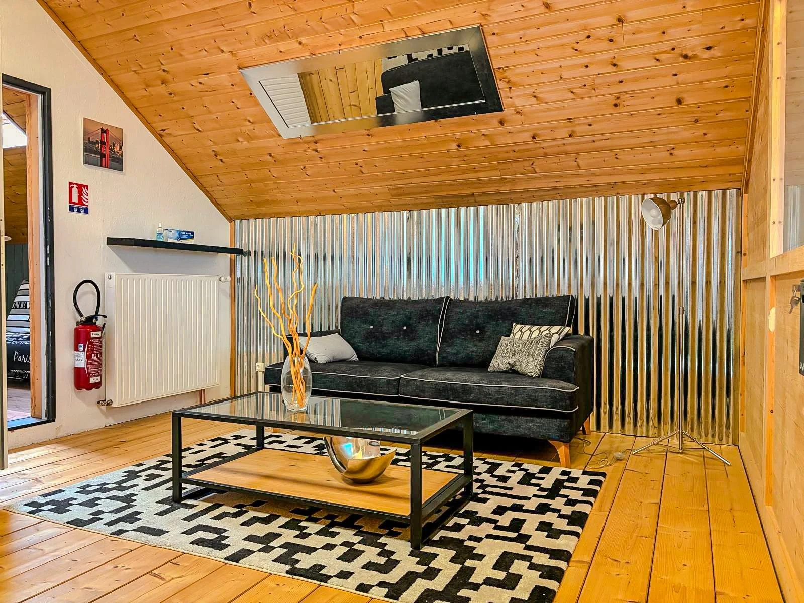 Salon avec un canapé noir, une table en verre avec un vase et des objets décoratifs, tapis noir et blanc, lampe sur le côté, mur en panneaux de bois et mur en métal, escalier visible à gauche.
