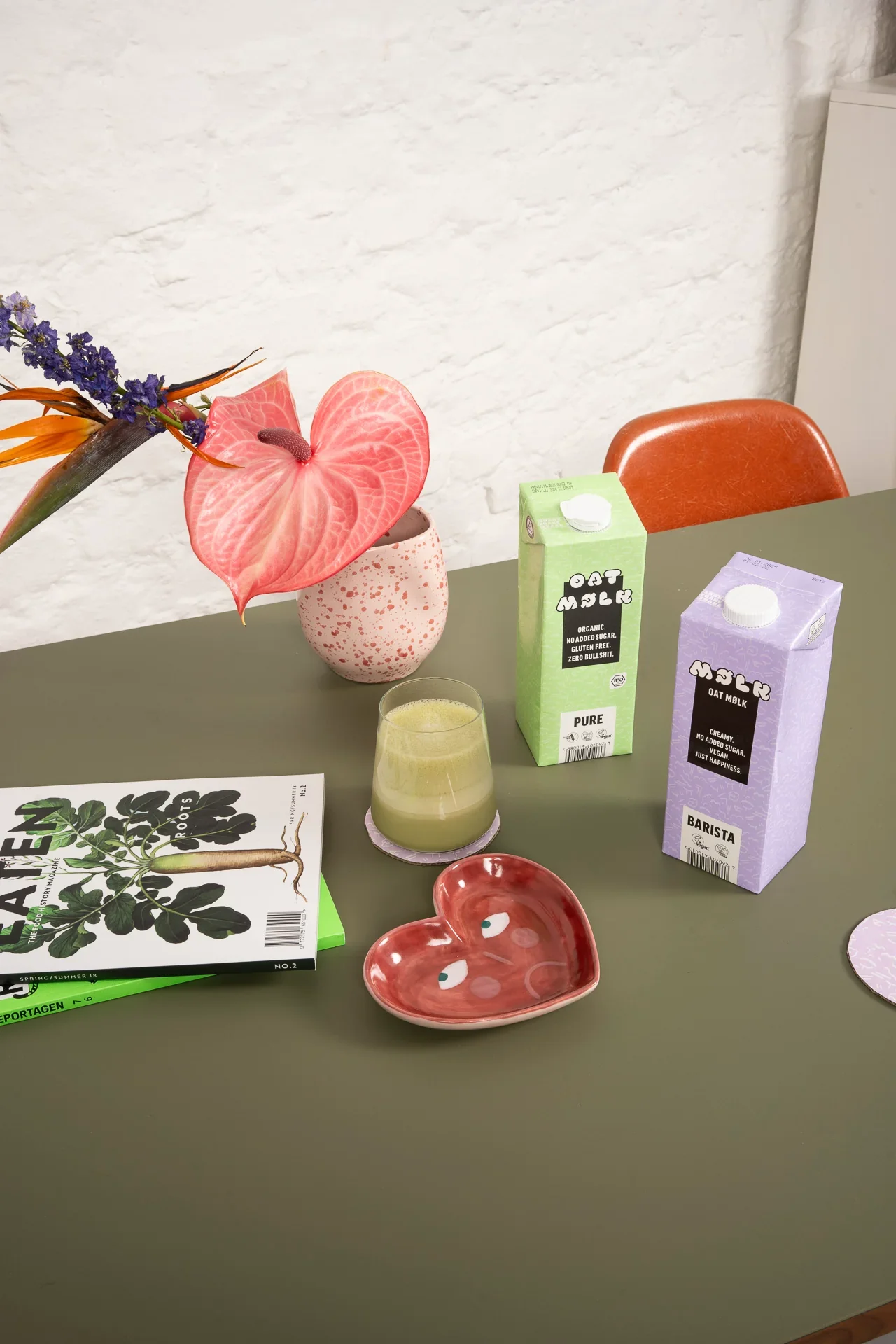 Tisch mit zwei Getränkekartons, einem Glas Saft, einer rosa Blumenvase mit bunten Blumen, einem Herz-Schälchen mit Augendekoration, Pflanzenbuch und Papptellern.