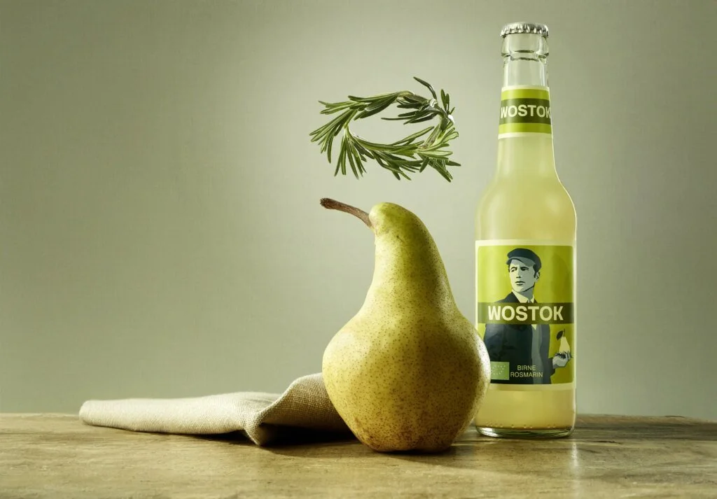 Eine Flasche Wostok Birne Rosmarin Limonade, eine reife Birne, ein Zweig Rosmarin, ein Stofftuch auf einem Holz Tisch. Hintergrund in einem neutralen Farbton.