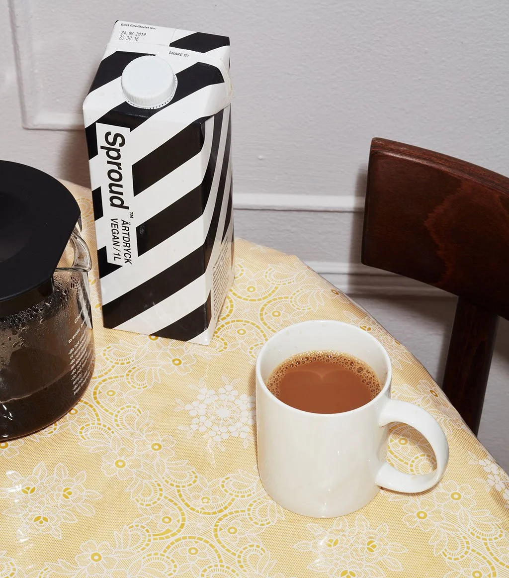 Auf einem Tisch steht eine weiße Tasse mit heißem Tee, eine Papiertüte mit veganem Spinat-Protein-Shake, eine Kanne mit dunkler Flüssigkeit, wahrscheinlich Kaffee, und ein Teil eines schwarzen Gegenstands. Der Tisch hat eine gelbe Decke mit weißen Mustern, und im Hintergrund ist eine dunkle Stuhllehne und eine weiße Wand.