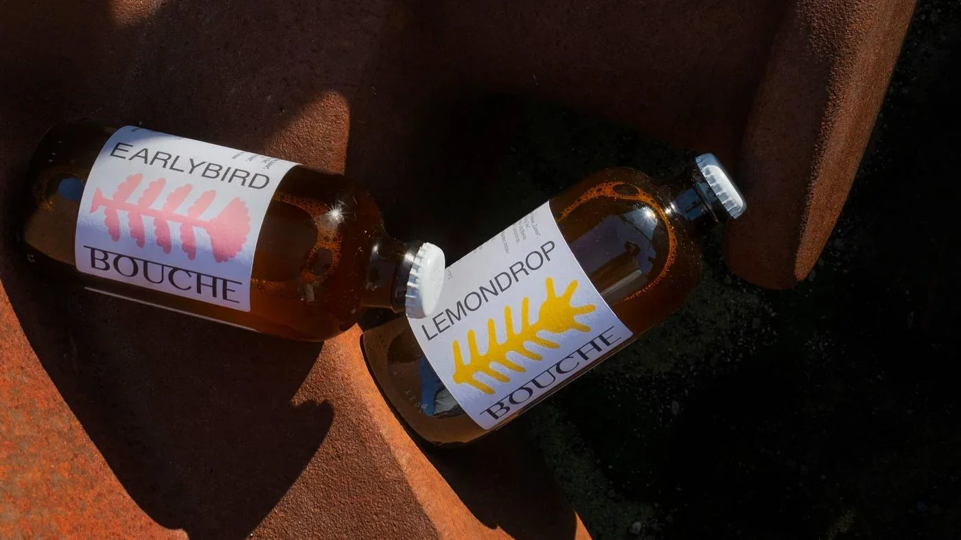 Zwei braune Flaschen mit labels, einer mit 'Earlybird Bouche' und der andere mit 'Lemondrop Bouche', liegen auf einer rustikalen Oberfläche im Sonnenlicht.