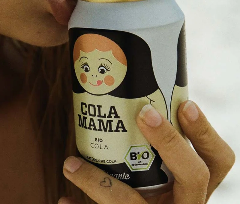 Eine Hand hält eine Dose mit einer Illustration eines Mädchens auf der Verpackung, das BIO Cola Produkt. Die Dose hat eine schwarze und cremefarbene Gestaltung.