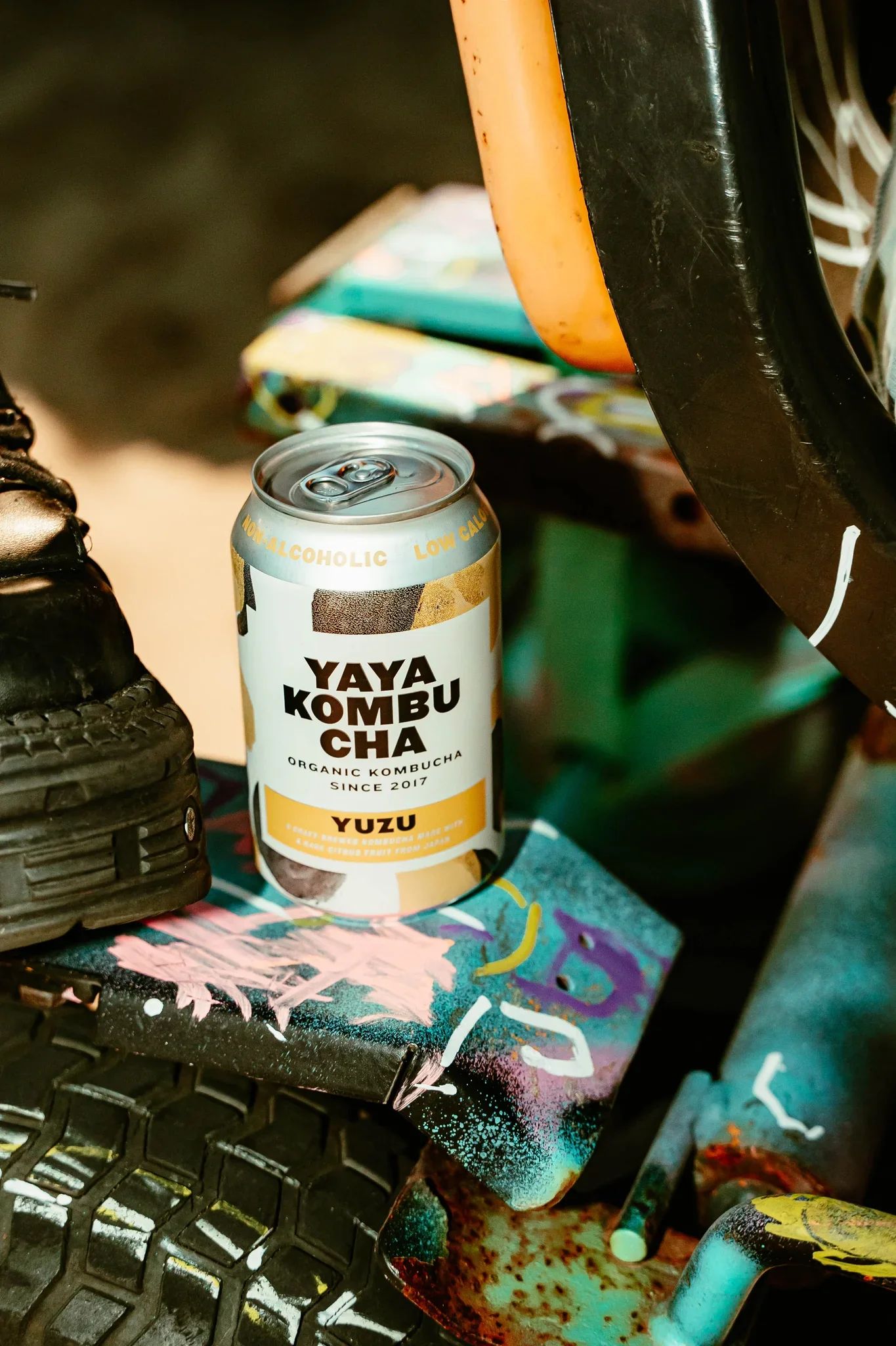 Alkoholische Getränkedose der Marke Yaya Kombucha, Geschmacksrichtung Yuzu, auf einem bunten, spritzenden Kunst- oder Graffiti-Kunstwerk. Umgebung mit schwarzem Schuh, Fahrradreifen und anderen Gegenständen.