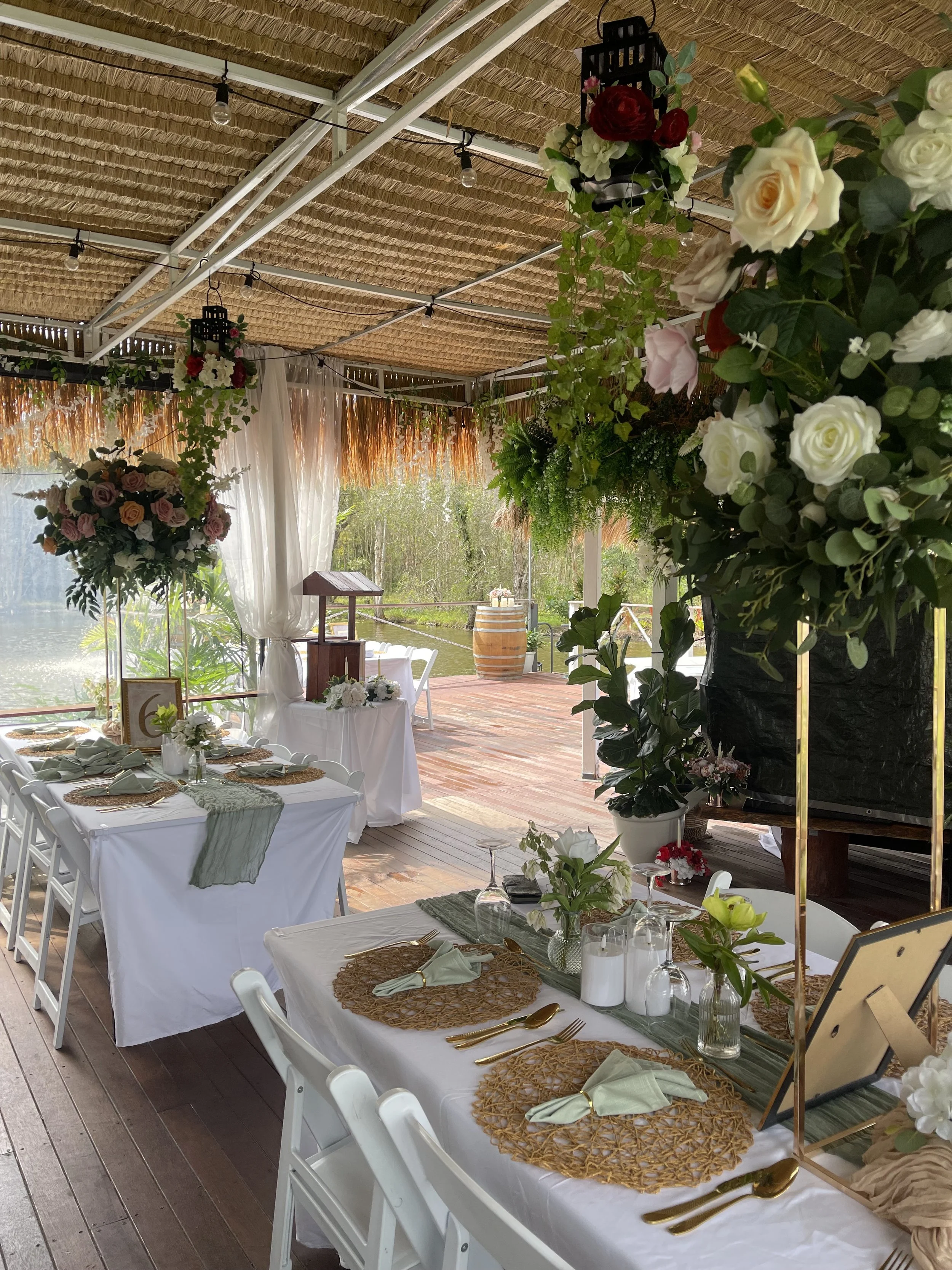 A decorated outdoor dining area with tables set for a celebration, featuring floral arrangements, greenery, and elegant tableware, with a thatched roof and view of a lake and nature in the background.