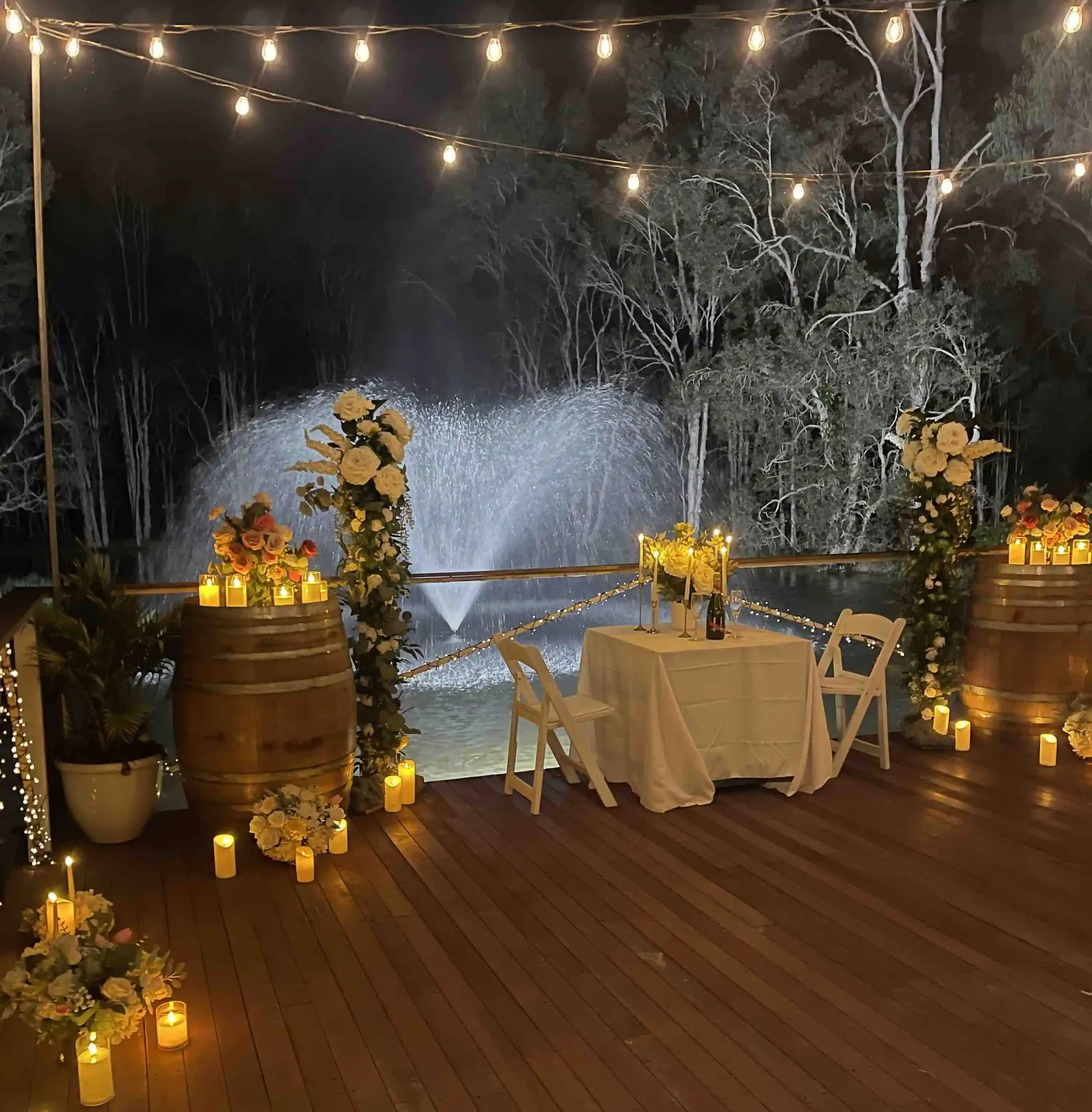 Nighttime outdoor setting with string lights overhead, flowers and candles decorating the scene, a waterfall in the background, and a table set for two with chairs.