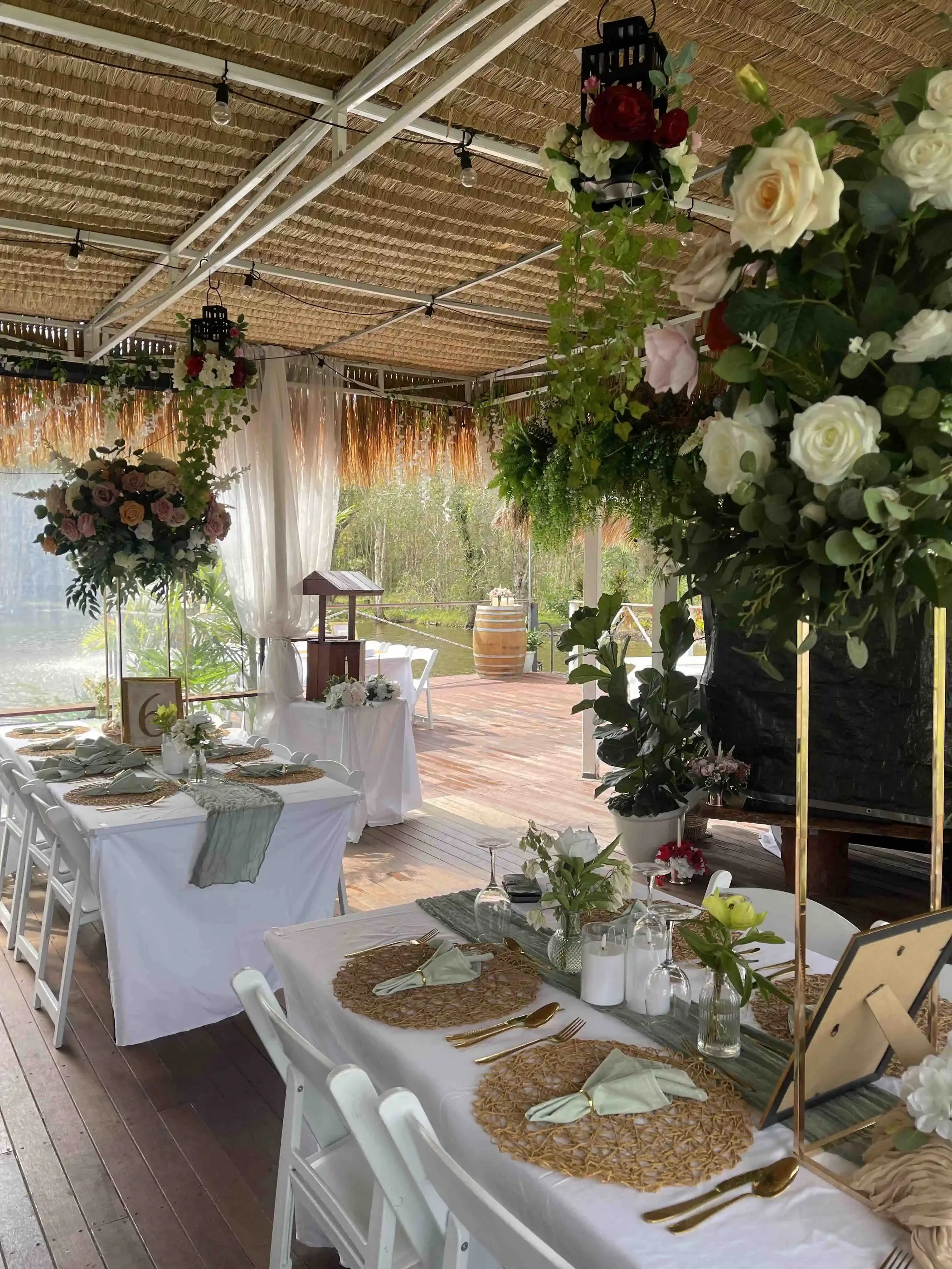 A decorated outdoor event space with a long table set with white tablecloths, gold utensils, green napkins, glassware, and floral centerpieces. The space has a thatched roof, hanging lights, and large floral arrangements with roses and greenery, with