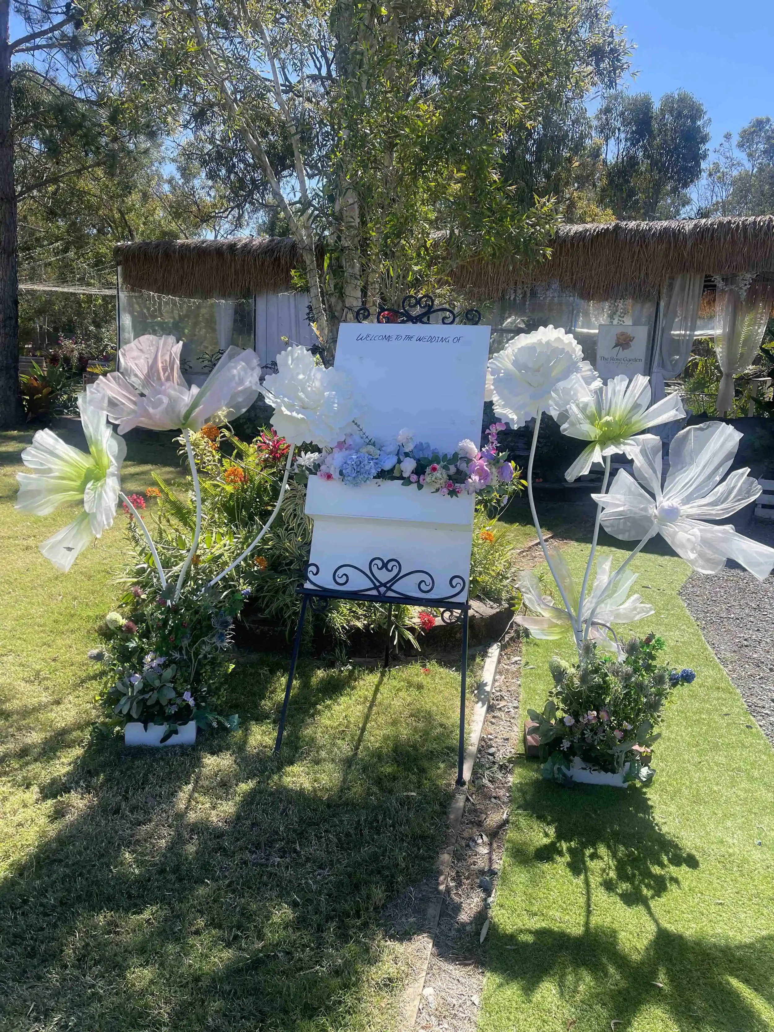 Outdoor wedding setup with white flowers, a sign that reads 'Welcome to the wedding of', and a thatched roof structure in the background under a clear blue sky.