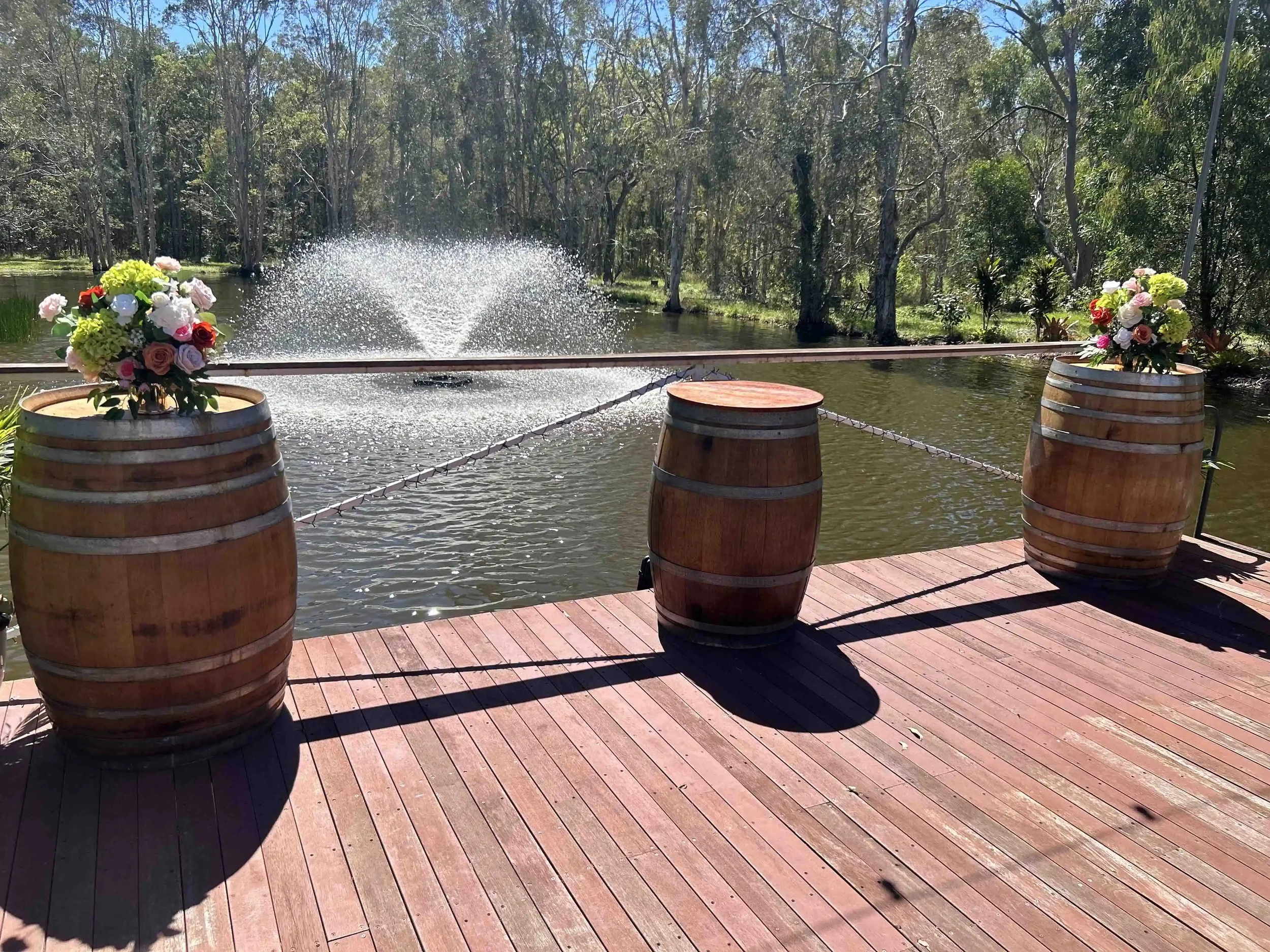 View of a dock by a river with trees in the background, decorated with two large flower arrangements in wooden barrels and a fountain in the water.