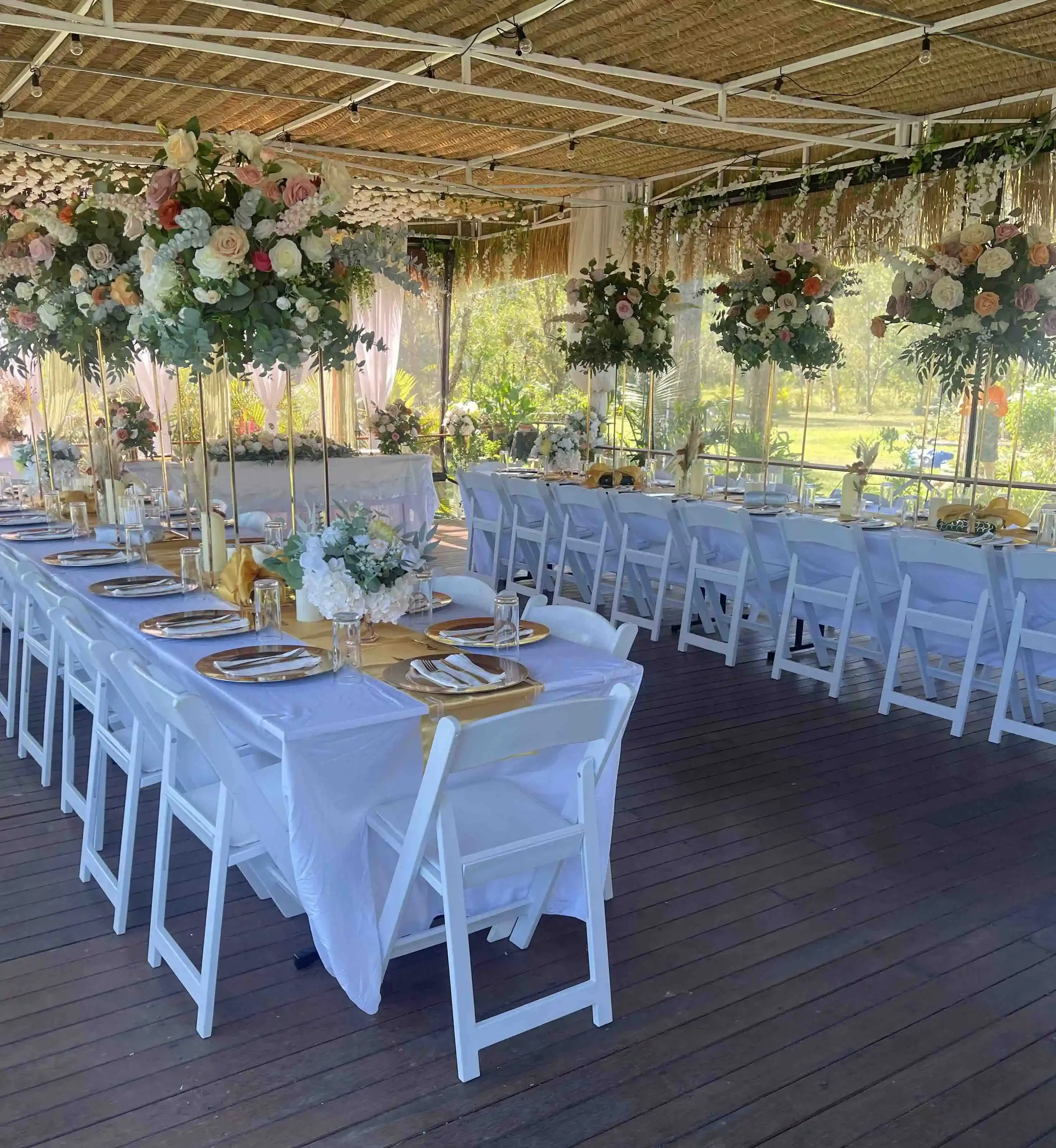Elegant outdoor wedding reception setup with long white tables decorated with floral centerpieces, place settings with gold chargers and white napkins, and white chairs. The venue has a thatched roof ceiling with string lights and lush greenery in th