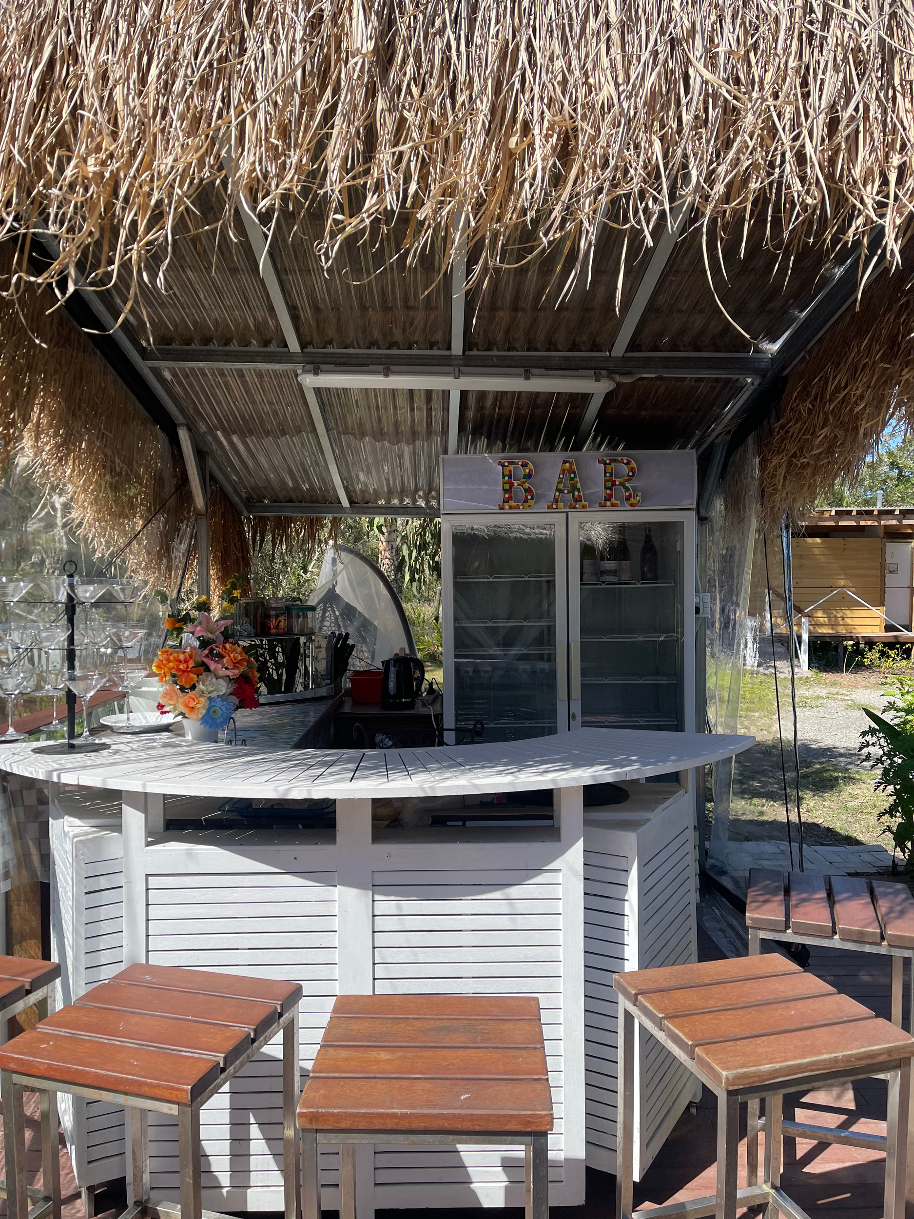 Outdoor bar area with a thatched roof and a glass-front fridge. Seating is available at the bar.