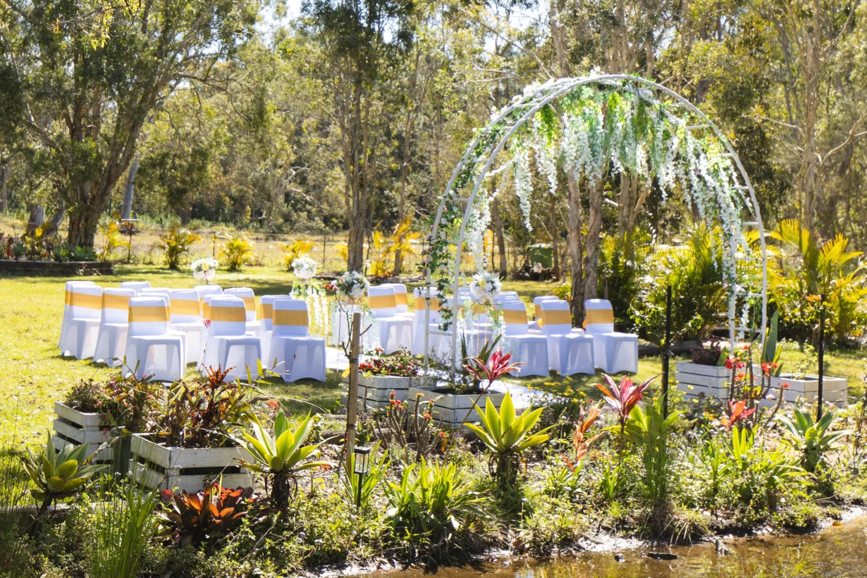 Outdoor wedding ceremony setup with white chairs decorated with gold ribbons, floral arrangements, and a white floral arch in a green garden with trees and a private lake.