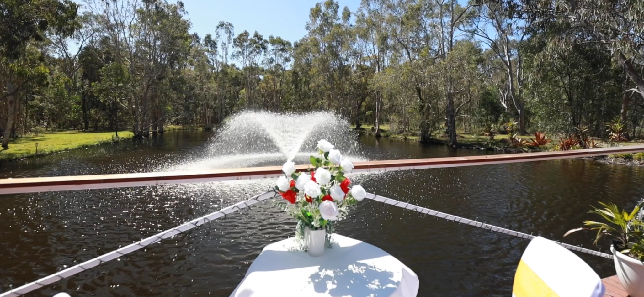 A boat decorated with a bouquet of white and red flowers on a table, floating on a river with a fountain spraying water, surrounded by green trees under a clear blue sky.