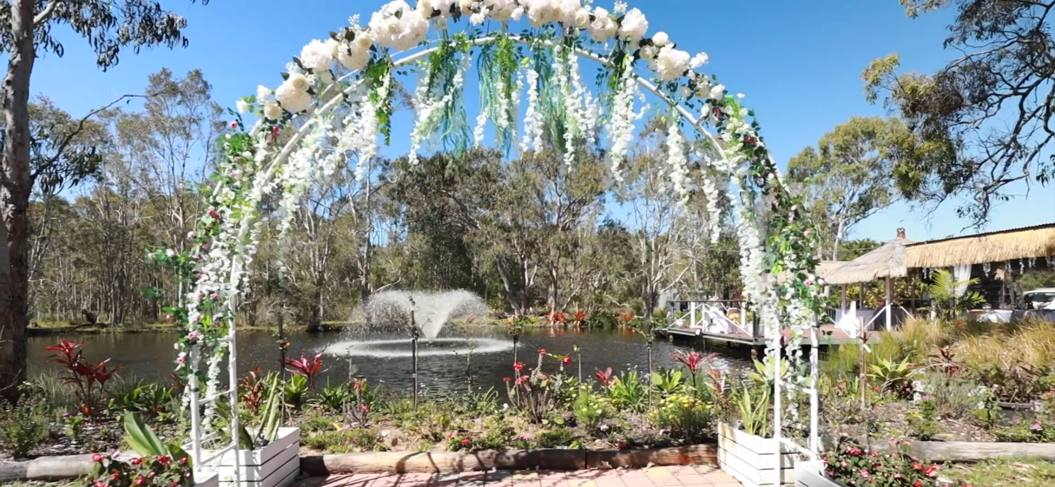 Decorated wedding arch with white flowers and greenery near a pond with a fountain, trees, and outdoor structures in background on a sunny day.