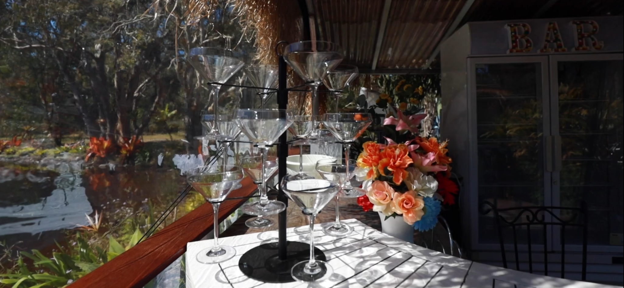 Glassware arranged on a white table with a black tiered stand holding empty cocktail glasses, next to a vase of colorful flowers, in a bright, outdoor setting with trees in the background.