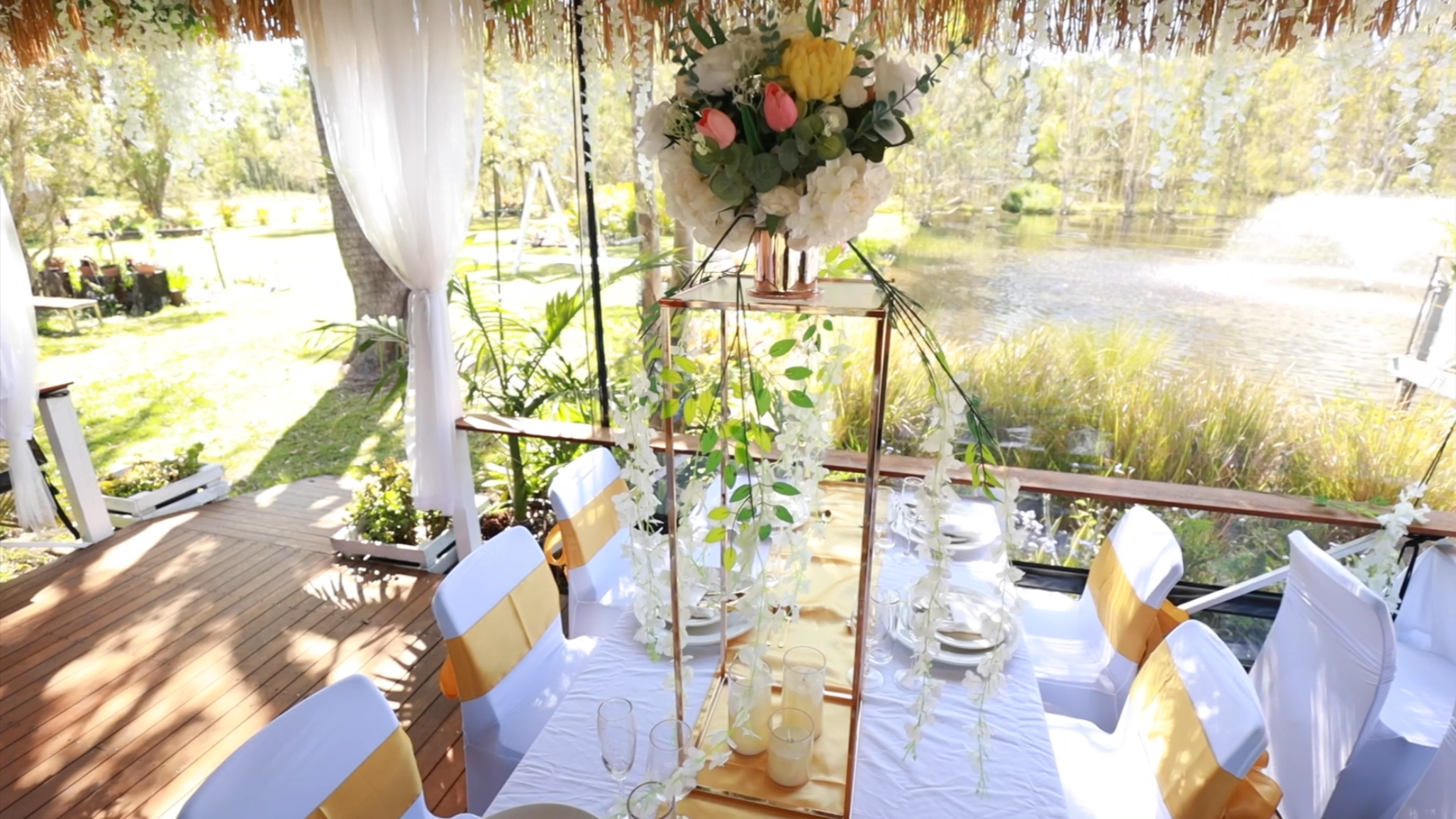 Decorated outdoor dining table with a floral centerpiece, surrounded by white chairs with yellow sashes, set near a lake with trees and grass in the background.