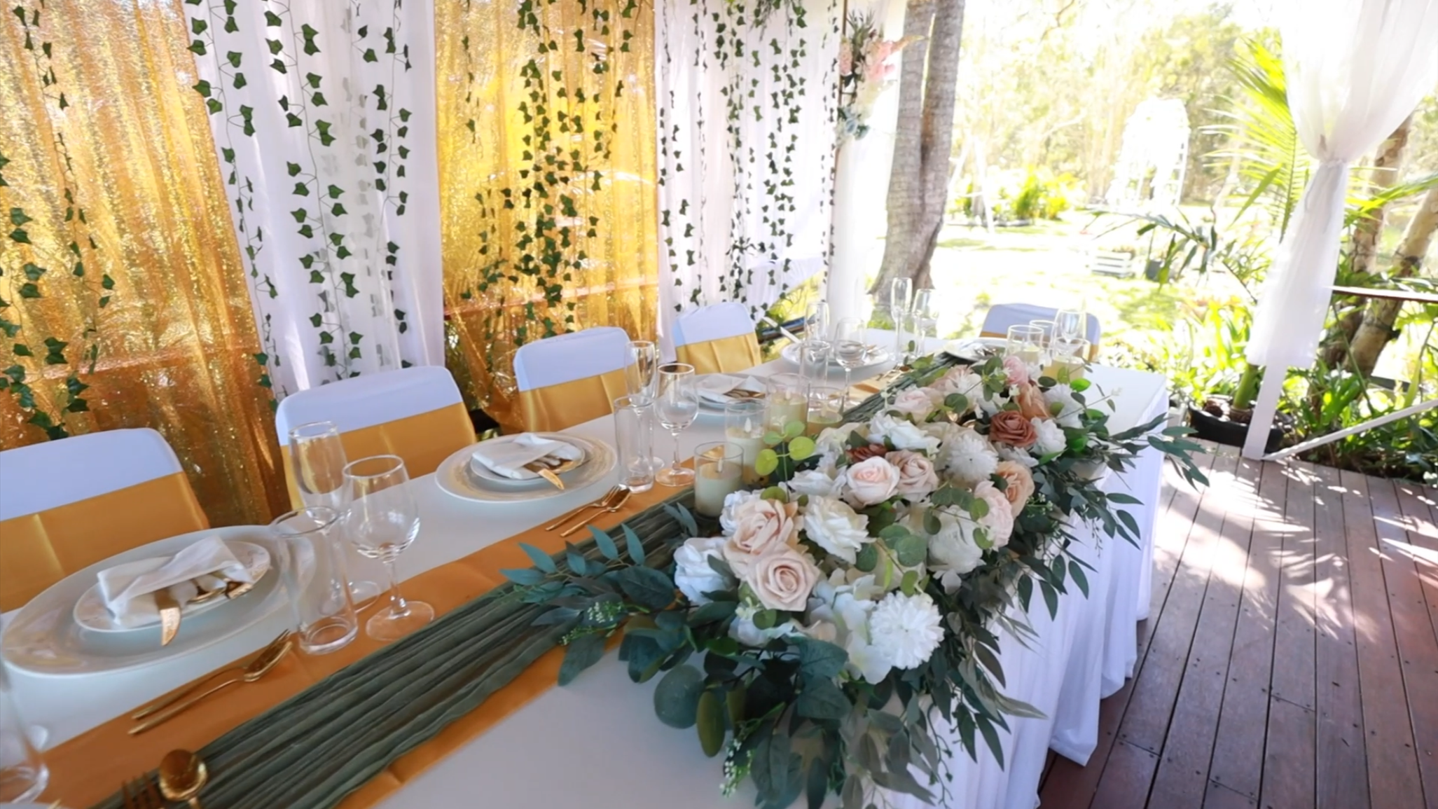 Decorated outdoor table set for a formal event with floral centerpiece, tableware, and gold accents, surrounded by greenery.
