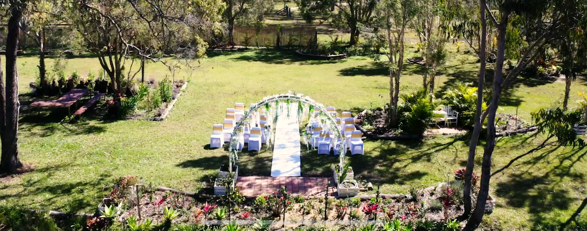 A backyard wedding setup with an arch decorated with flowers and white chairs arranged on either side of a aisle on grass, surrounded by trees and plants.