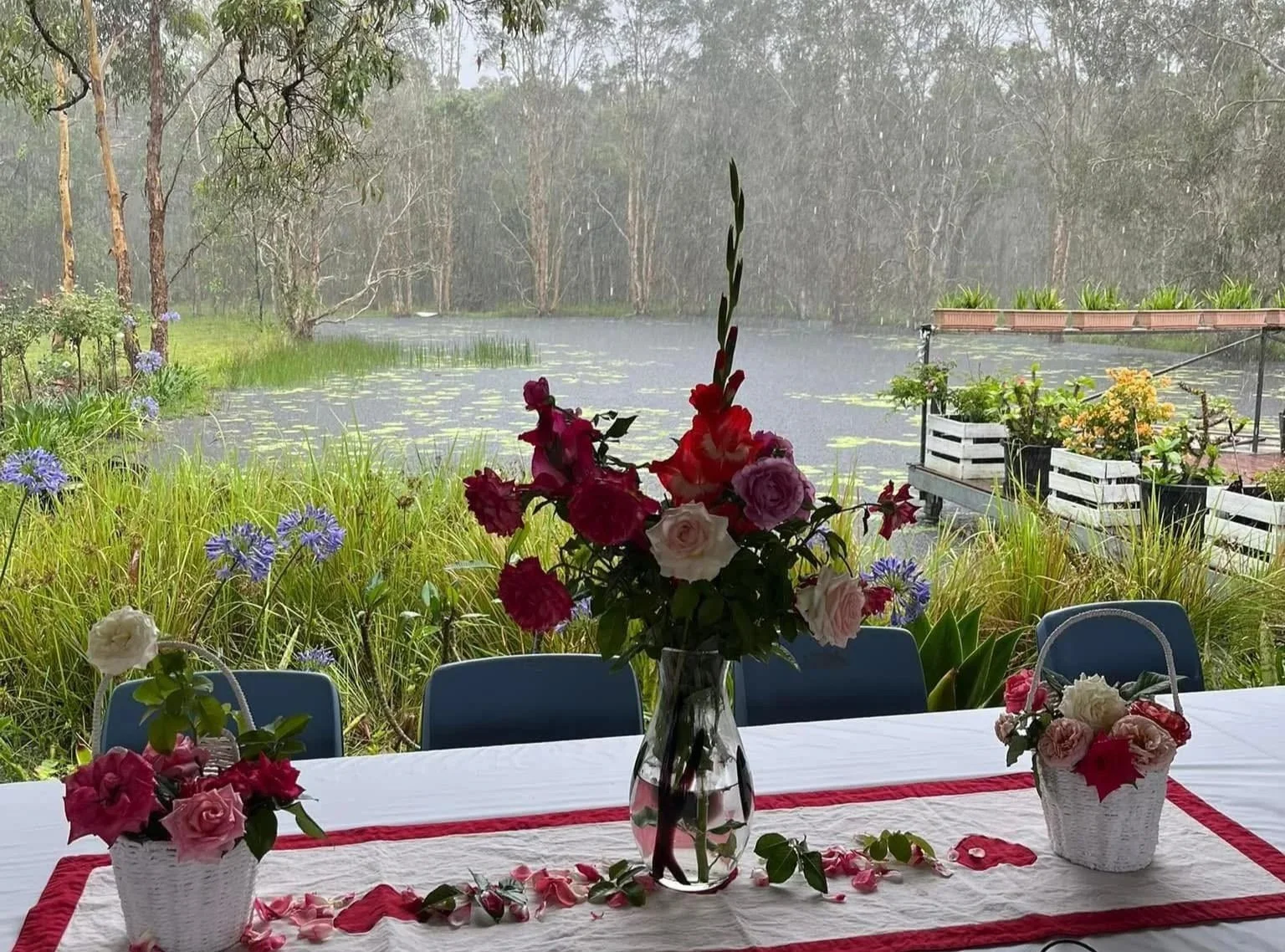 Table with a white tablecloth and red accents, decorated with pink, white, and purple flowers in baskets and a tall glass vase, set outdoors with a lake, trees, and rain in the background.