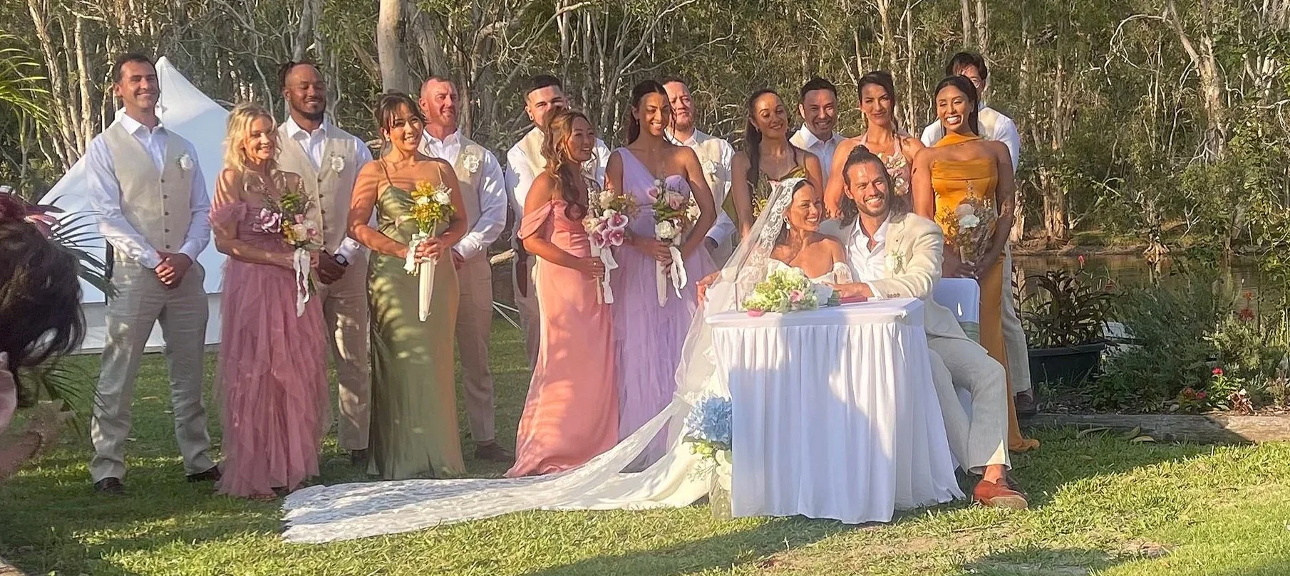 A wedding celebration outdoors by a private lake, with a bride and groom sitting at a table, surrounded by wedding party members holding bouquets and dressed in pastel and vibrant colours.