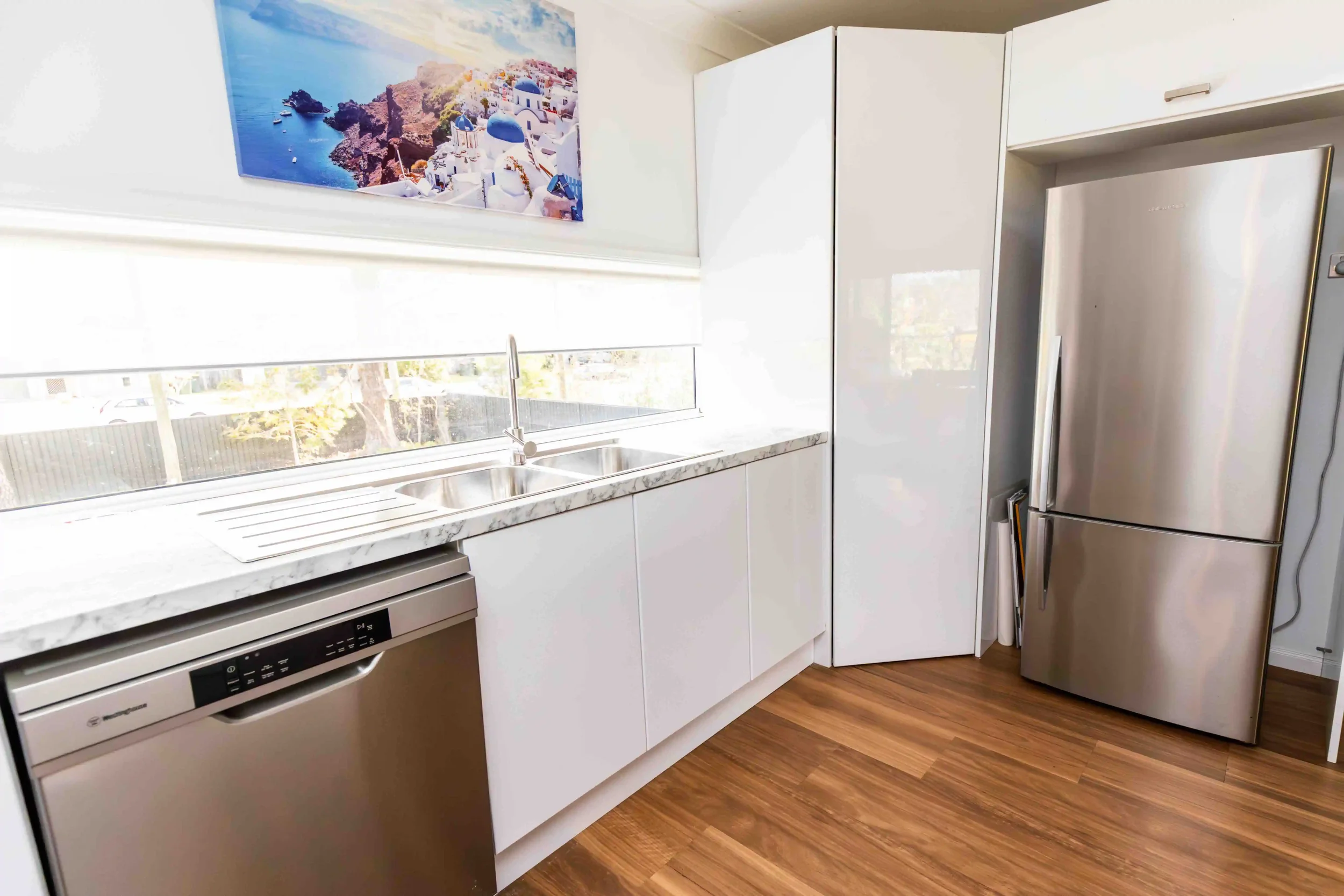 Modern kitchen with white cabinets, marble countertop, stainless steel refrigerator, dishwasher, and a window with a scenic view of a coastal town with white buildings and blue domes.