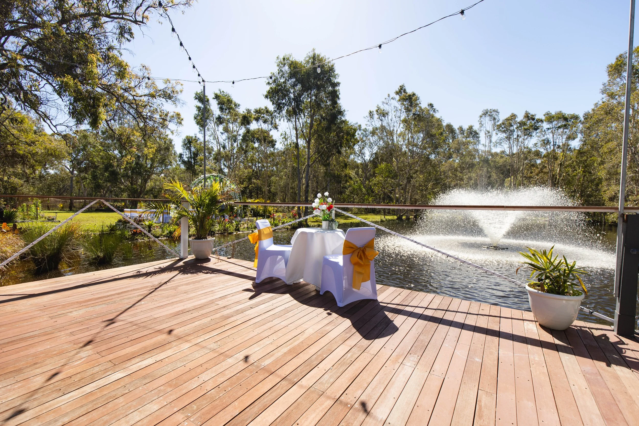 A decorated outdoor deck overlooking a pond with a fountain, featuring a small round table with a floral centerpiece and two chairs with white covers and gold ribbons, potted plants, and string lights overhead.