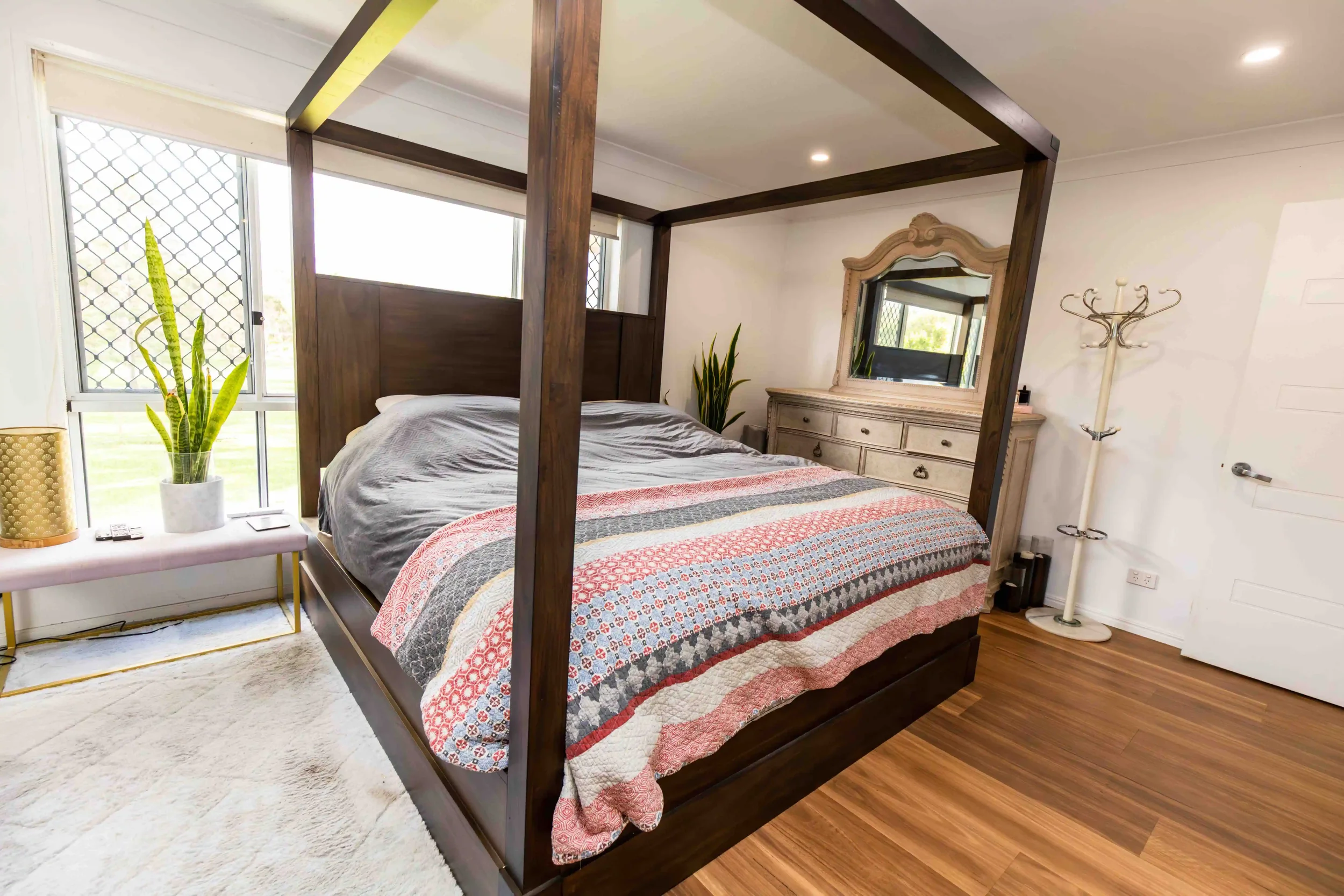 A bedroom with a four-poster wooden bed, a vintage-style dresser with a mirror, potted plants, a coat rack, a side table with a plant, and large windows with security screens.