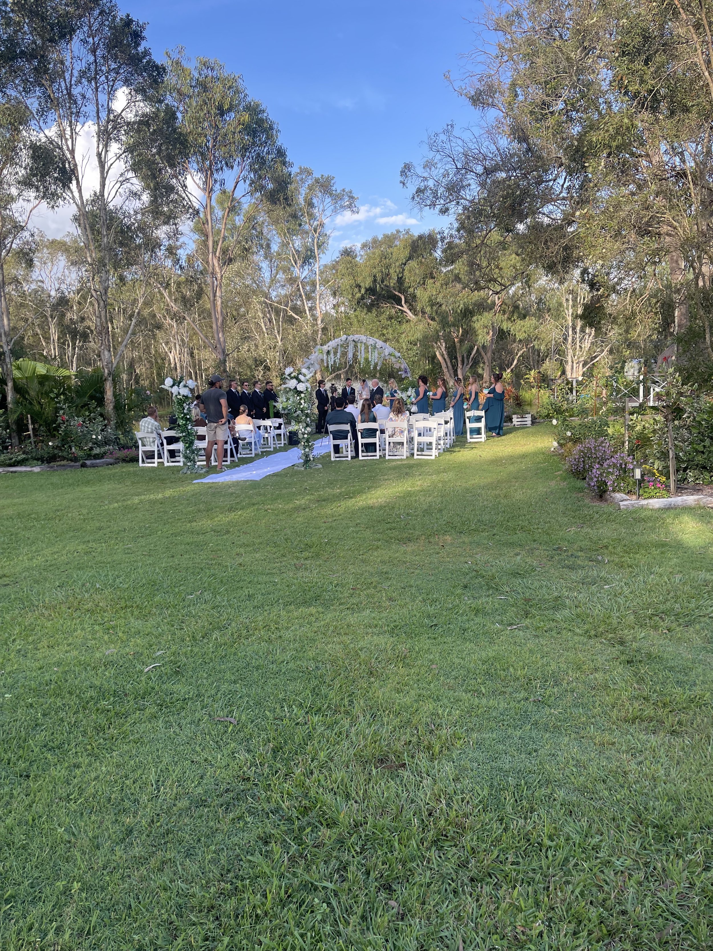 Outdoor wedding ceremony setup on a lush green lawn with chairs and floral decorations under a canopy of trees and blue sky.