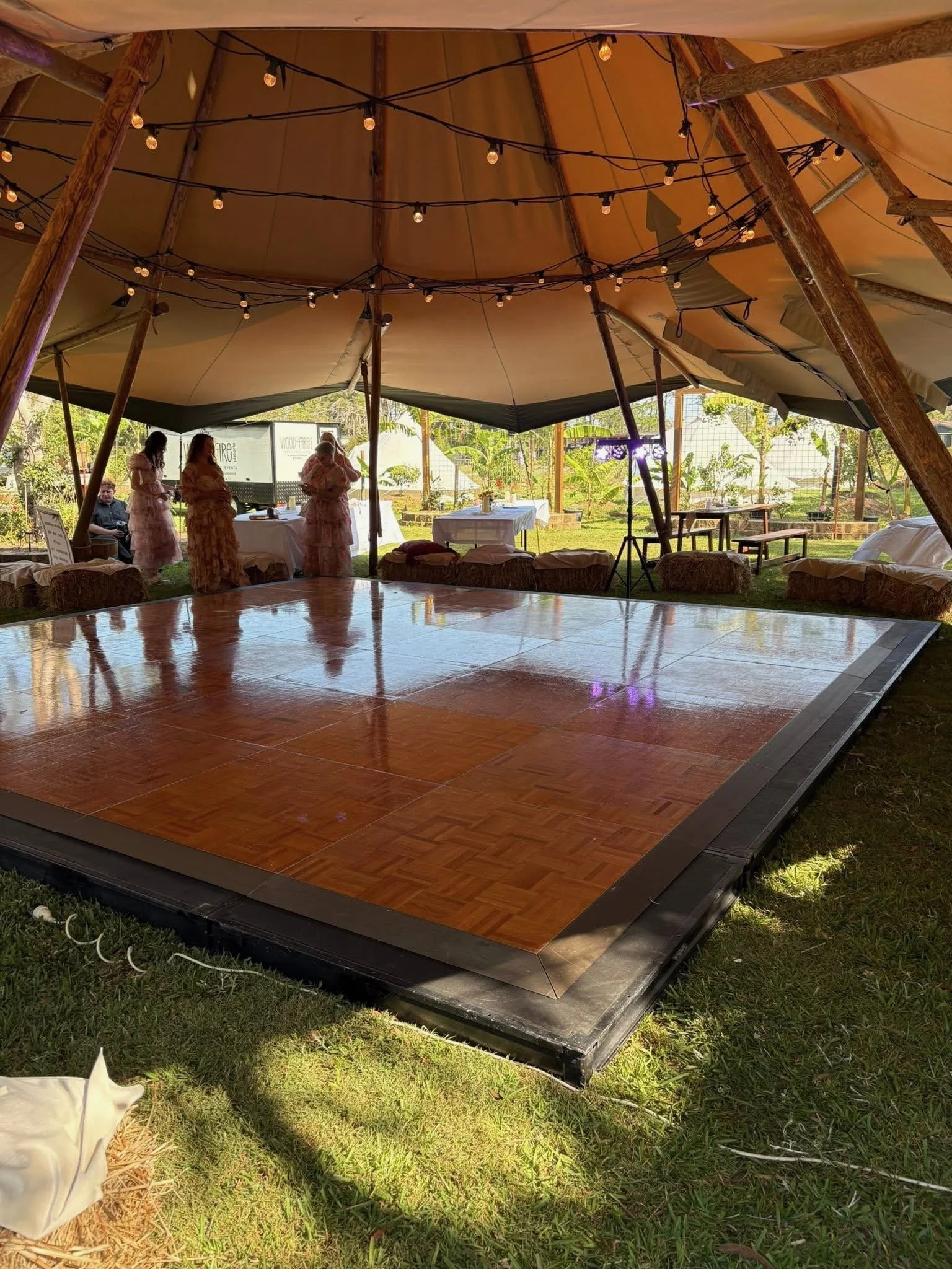 Indoor event space with a wooden dance floor under a large canvas tent, decorated with string lights. Several people in pink dresses are standing near a table in the background.