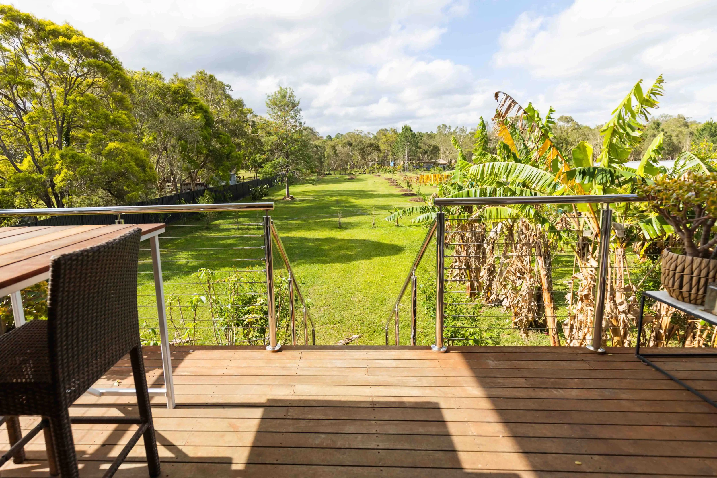 View from a wooden porch with outdoor furniture, overlooking a green grassy yard with trees and banana plants, under a partly cloudy sky.