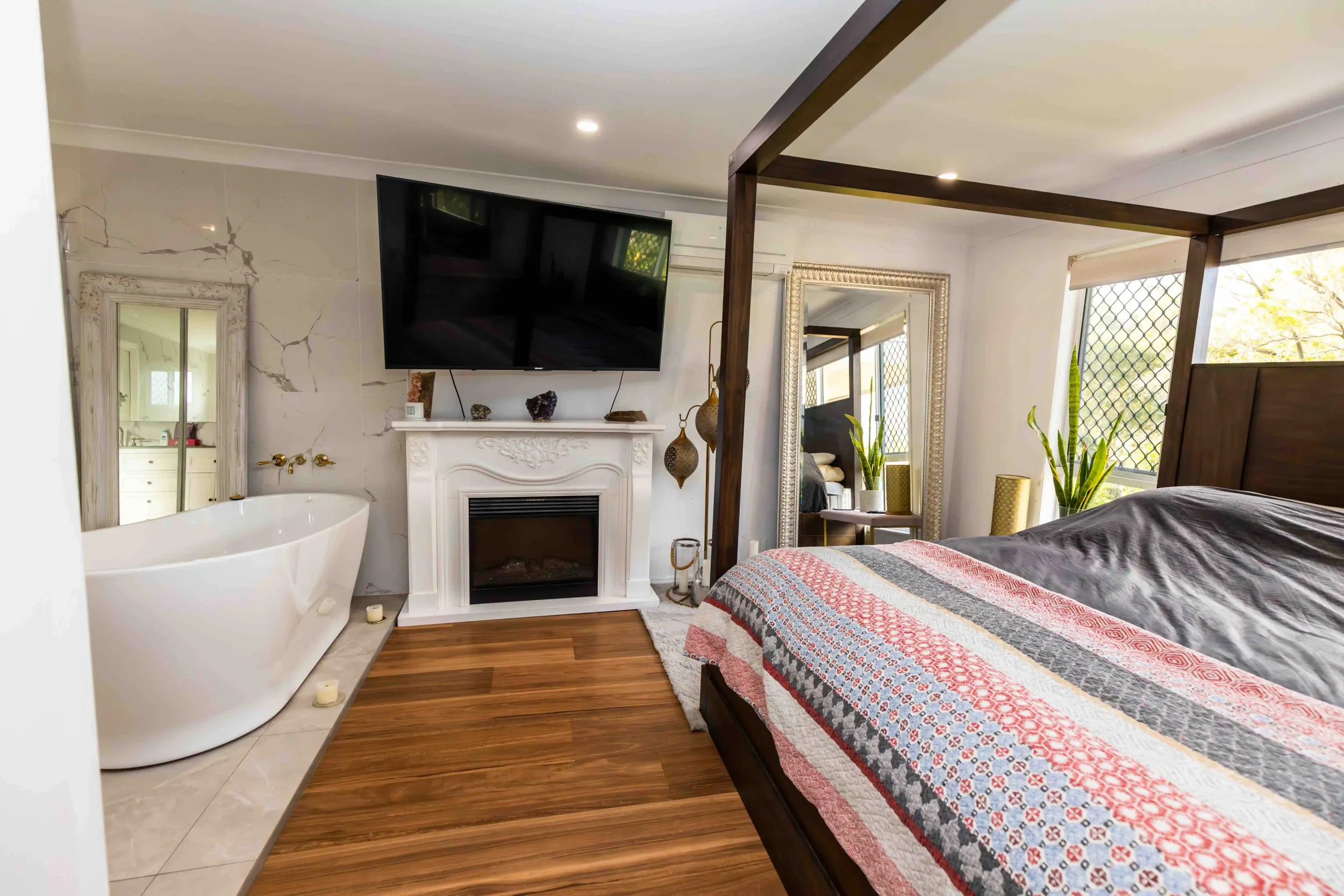 A cozy bedroom with a large bed covered in a colorful patchwork quilt, a white fireplace with decorative items, a flat-screen TV mounted above the fireplace, a tall mirror with a decorative frame, a potted plant, and a freestanding mirror reflecting part of the room. The room features wood flooring, large windows with security screens, and a mix of modern and vintage decor.