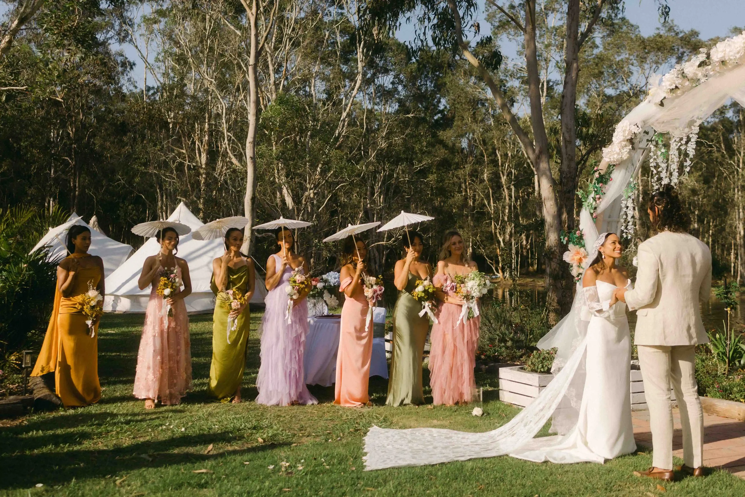 Women in colorful dresses holding bouquets, standing in a line outdoors, with a bride and groom at the altar, during a wedding ceremony in a wooded area.